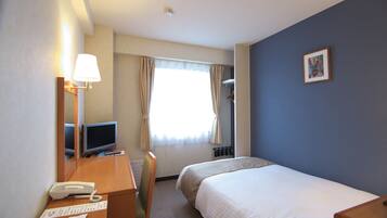 A hotel room with a bed, a desk with a telephone and a computer, a chair, a window with curtains, and a framed picture on the wall.