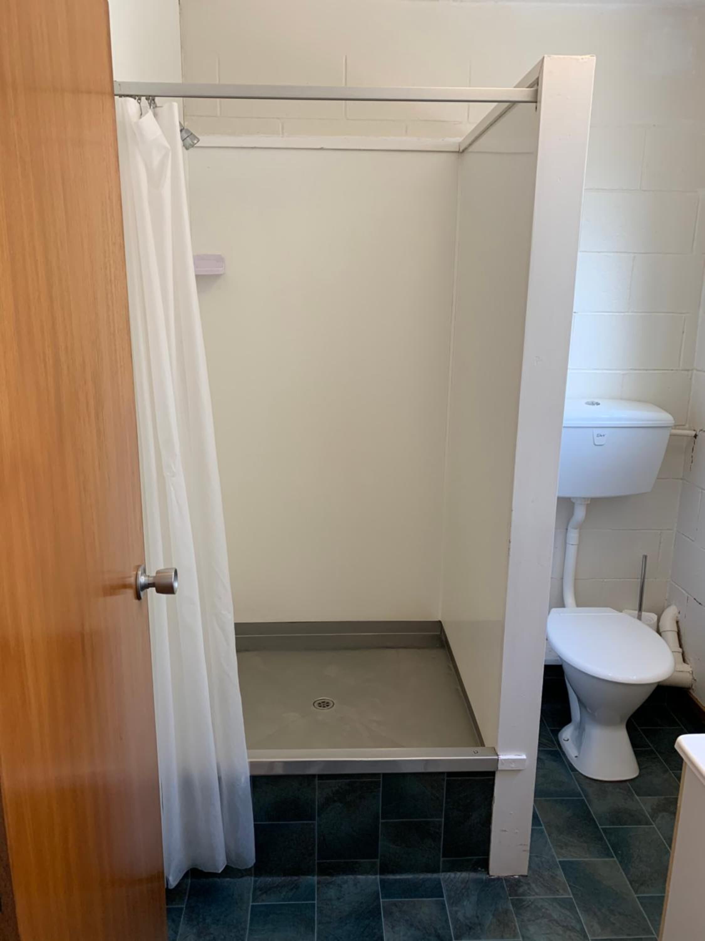 Um chuveiro pequeno e fechado com cortina branca, vaso sanitário branco e chão revestido de azulejos.