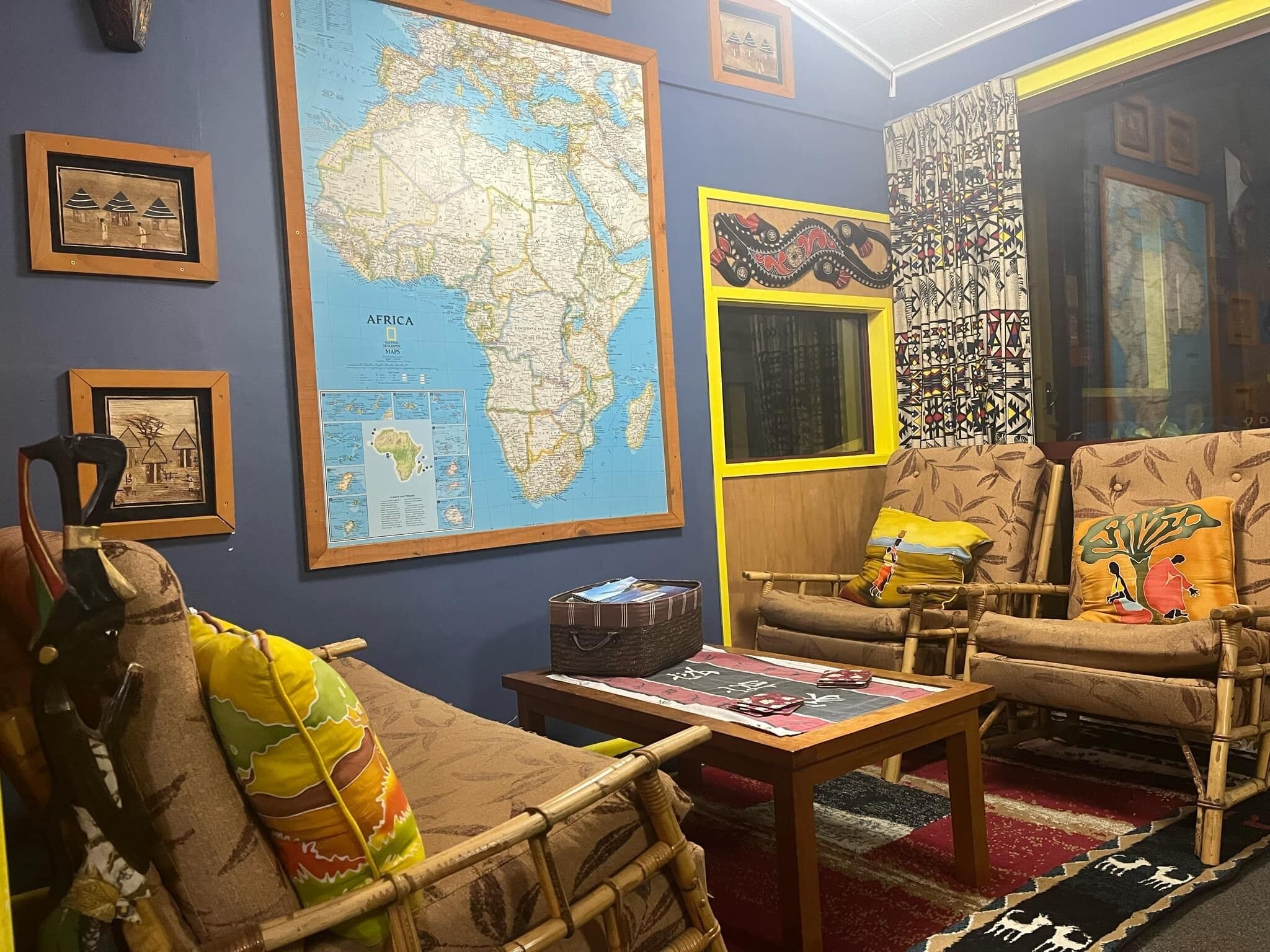 A room with a large map of Africa, two wicker chairs with cushions, a small table with a magazine, and a decorative wall piece.