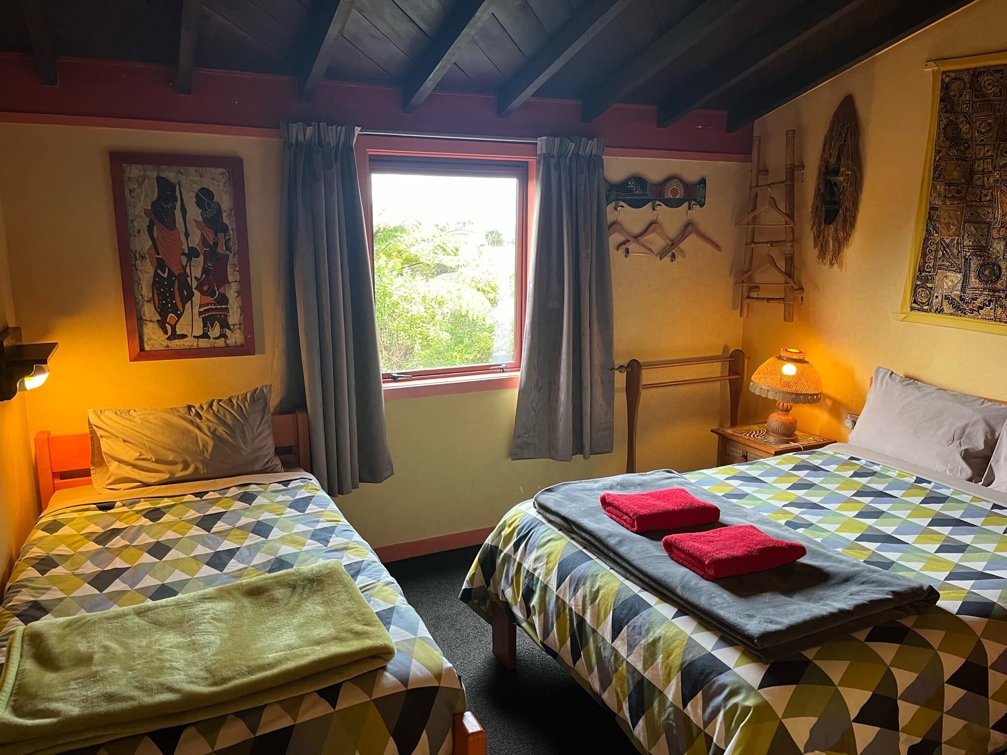 A room with two beds, each with a patterned bedspread, a window with curtains, a wall painting, a wooden headboard, and a nightstand with a lamp.