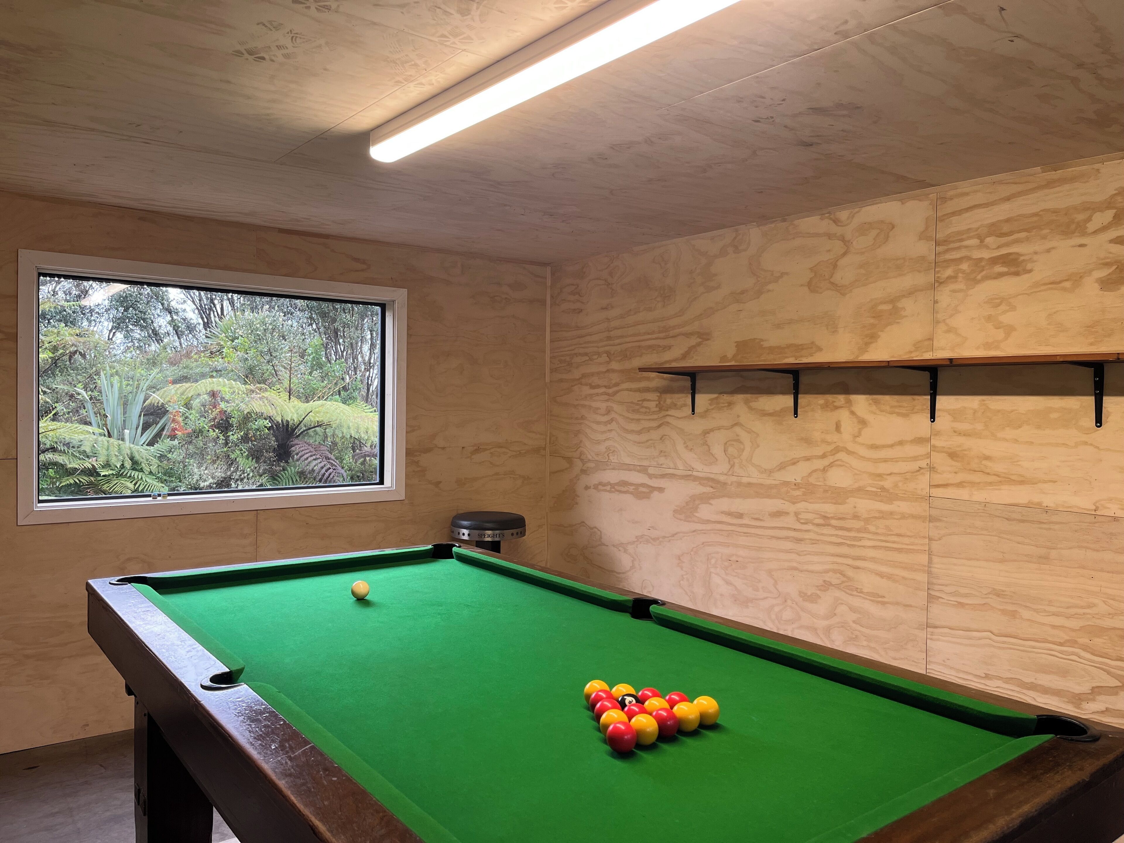 Une table de billard avec des billes, une fenêtre donnant sur un paysage verdoyant et une étagère avec un porte-queue de billard.