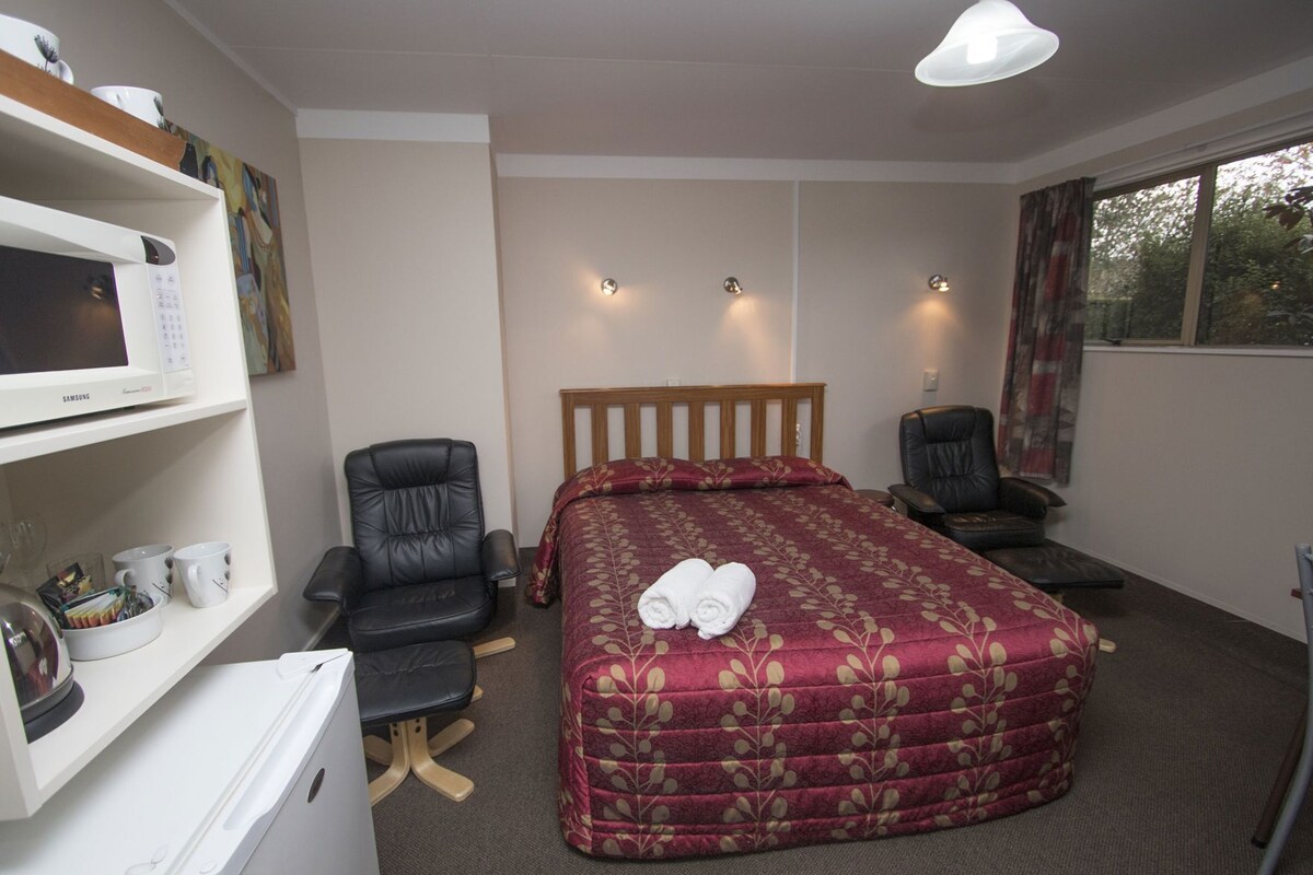 A hotel room with a bed, two chairs, a desk with a microwave, and a window.