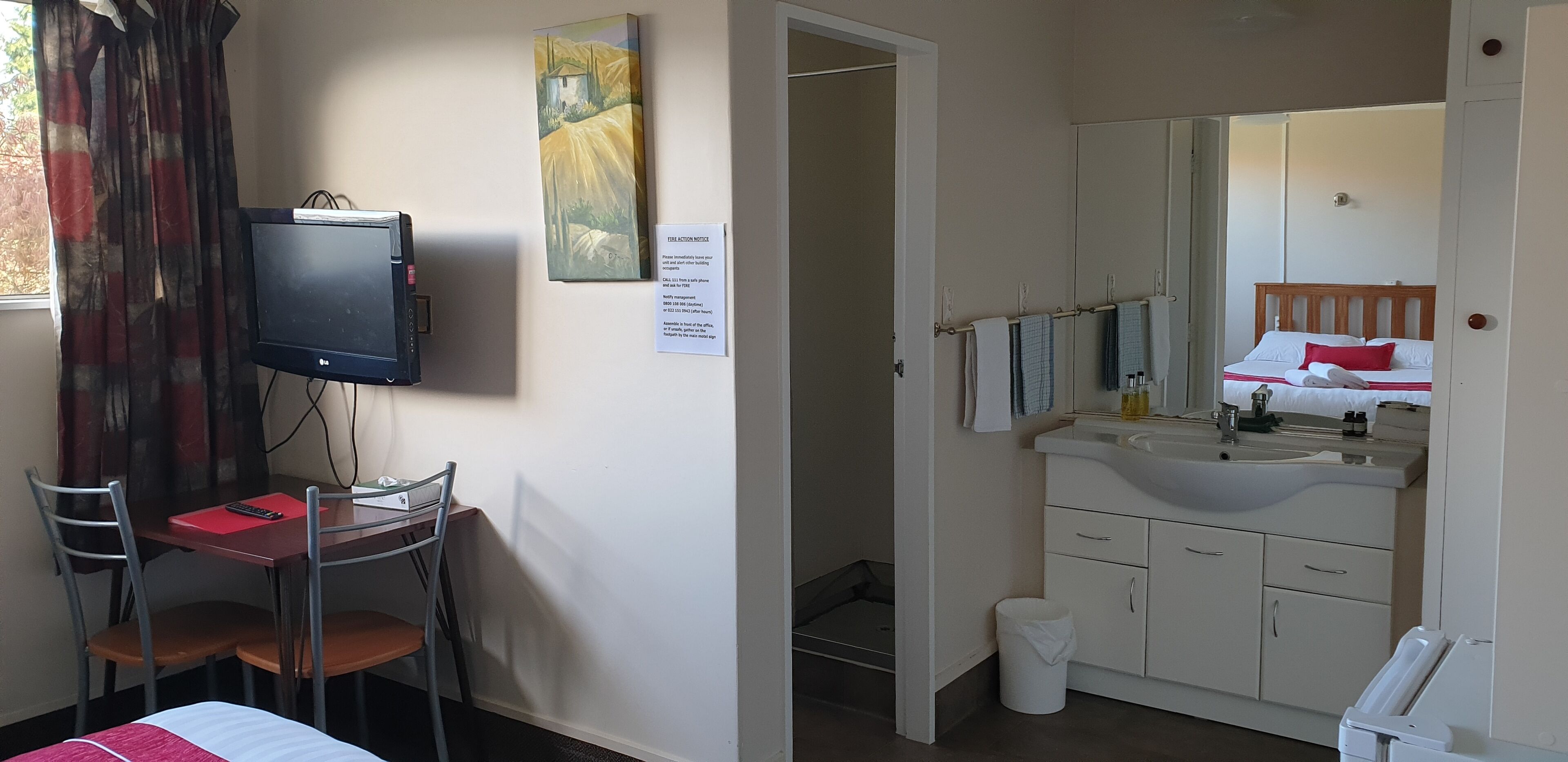 A hotel room with a bed, a desk with a chair, a television, a bathroom with a sink and mirror, and a painting on the wall.