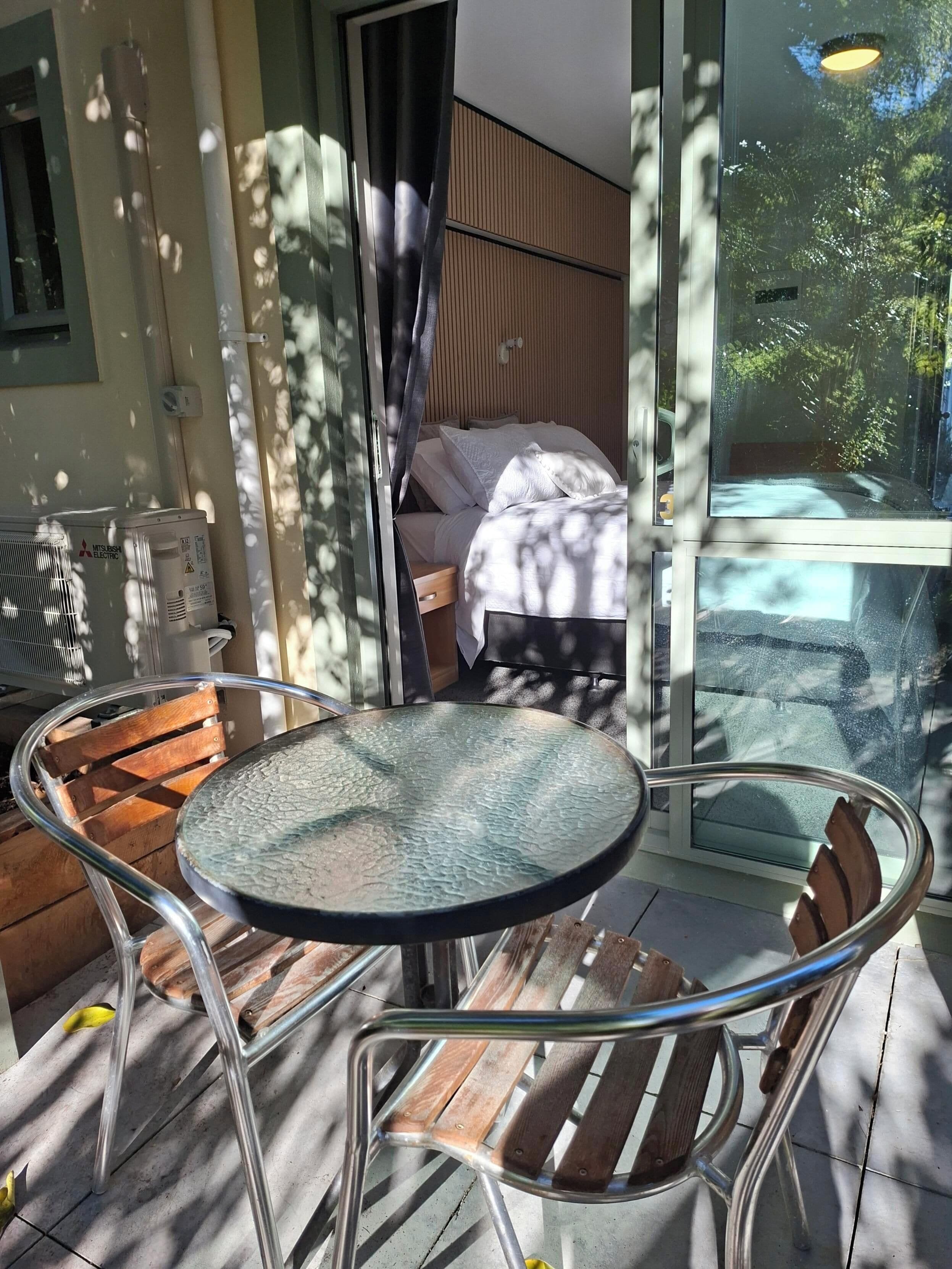 A glass table with chairs on a balcony, a bed with white bedding, and a view of trees through the glass.