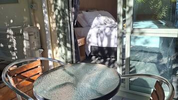 A glass table with chairs on a balcony, a bed with white bedding, and a view of trees through the glass.