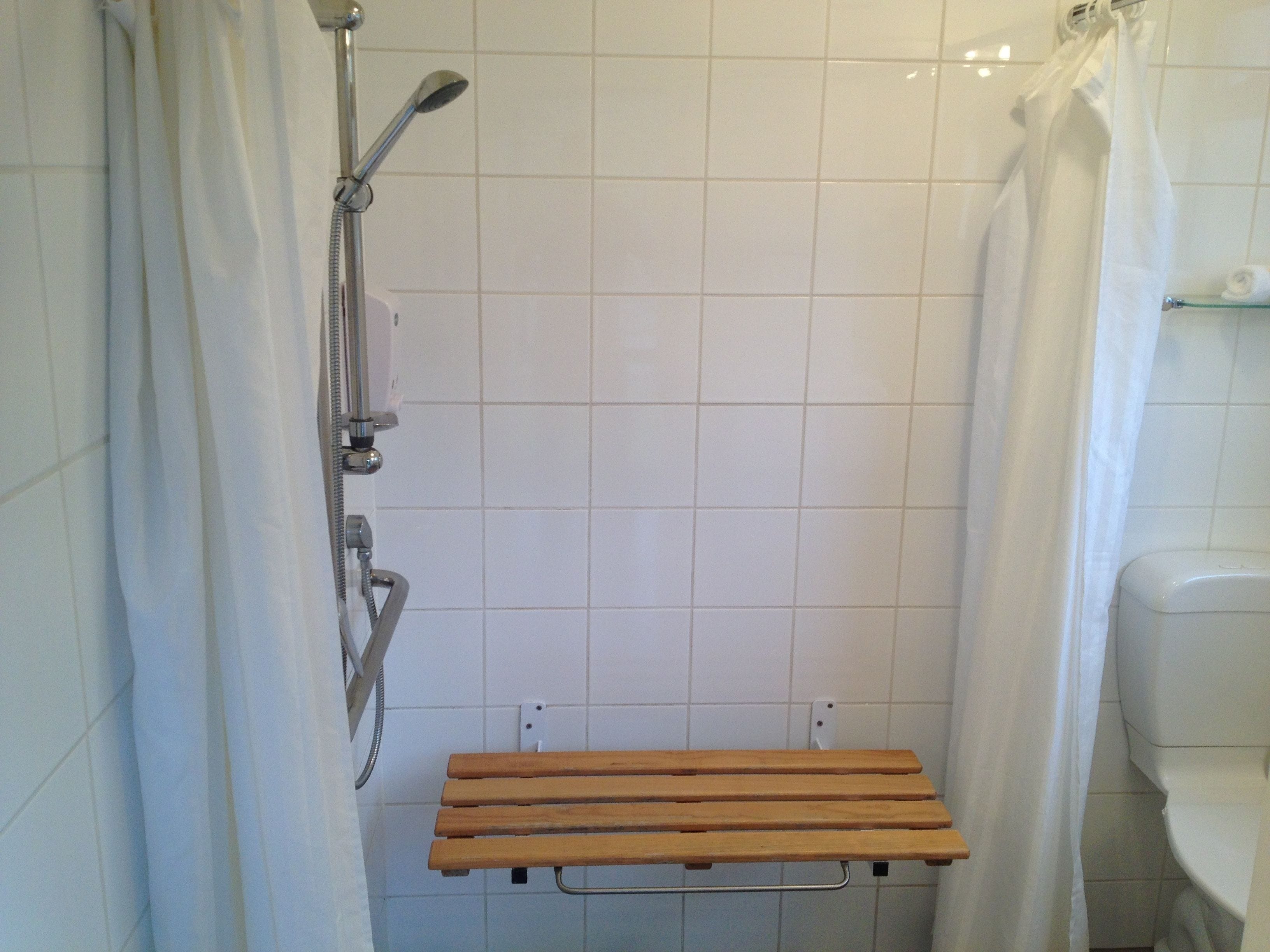 A shower area with a wooden bench, white tiled walls, and a glass shelf above the toilet.
