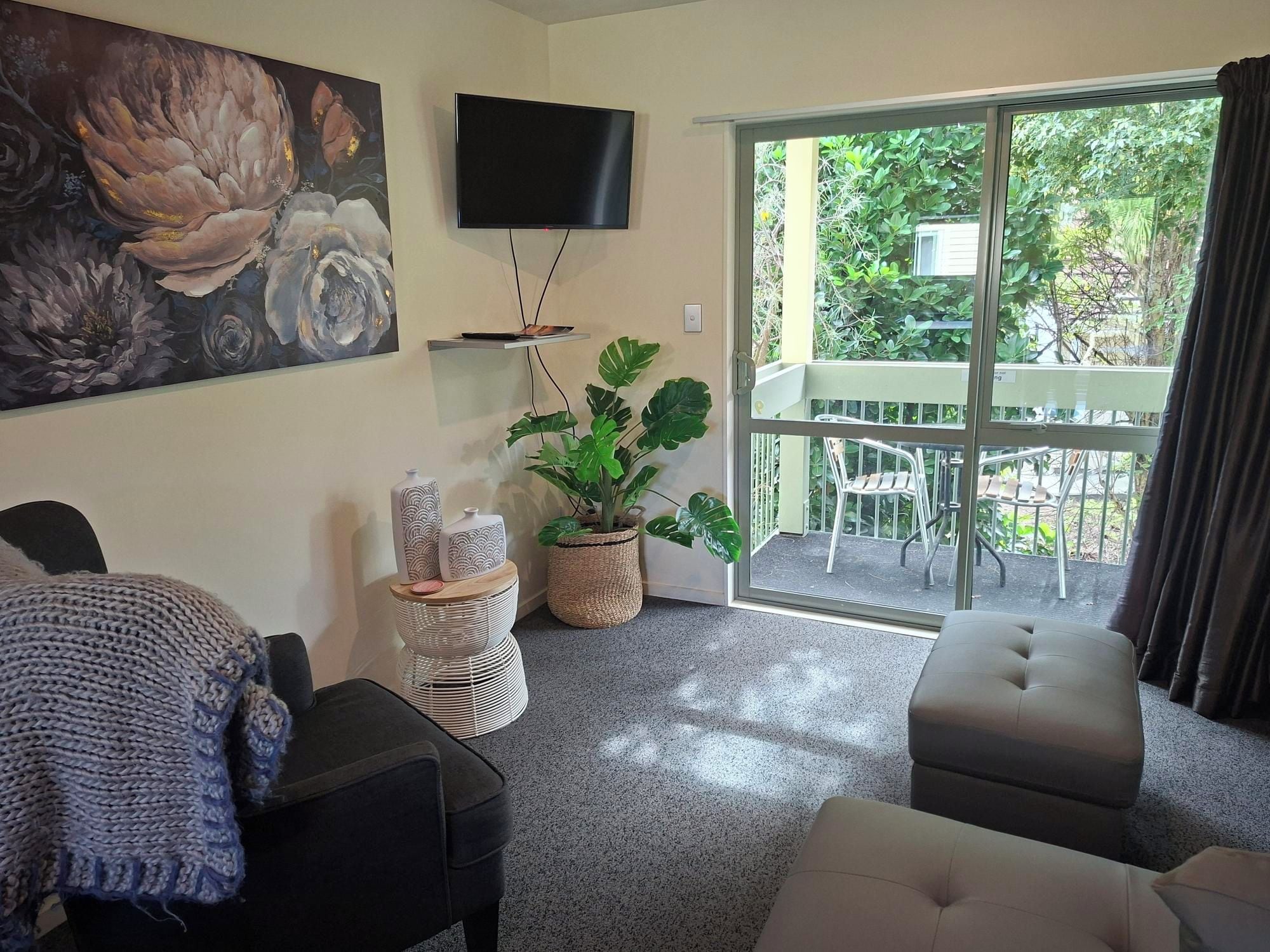 A living room with a flat-screen TV, a large floral painting, a potted plant, and a balcony with outdoor furniture.