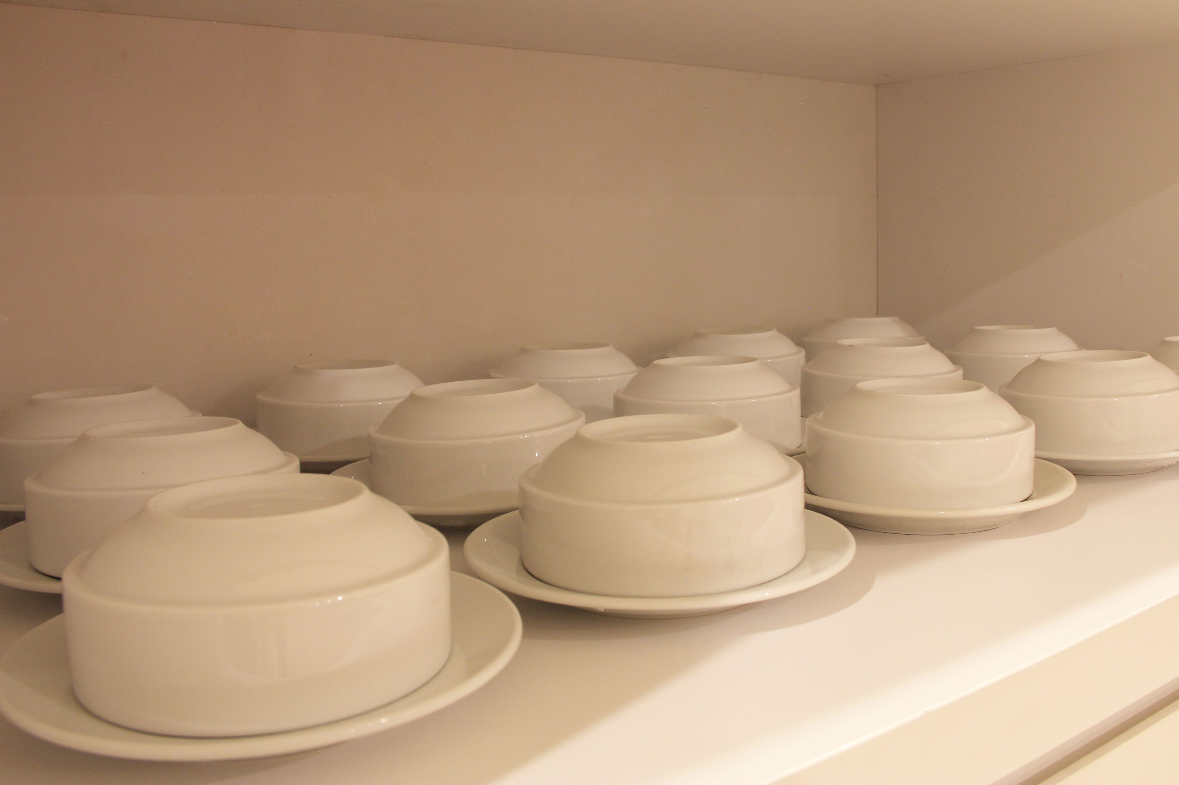 Una hilera de cuencos blancos de cerámica sobre platos blancos, dispuestos ordenadamente sobre una superficie blanca.