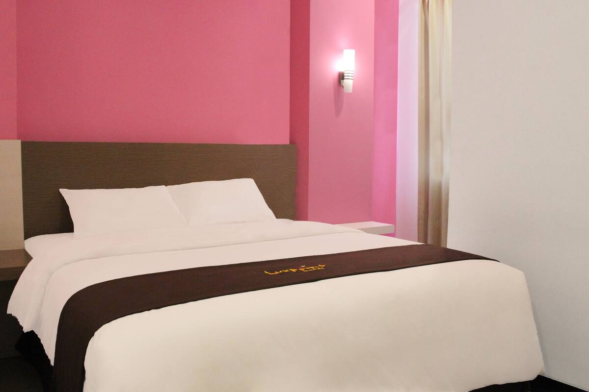 A hotel room with a bed, a nightstand, and a wall-mounted light.