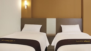 Quarto de hotel com duas camas, uma mesa de cabeceira, uma luminária fixada na parede e uma parede lisa.