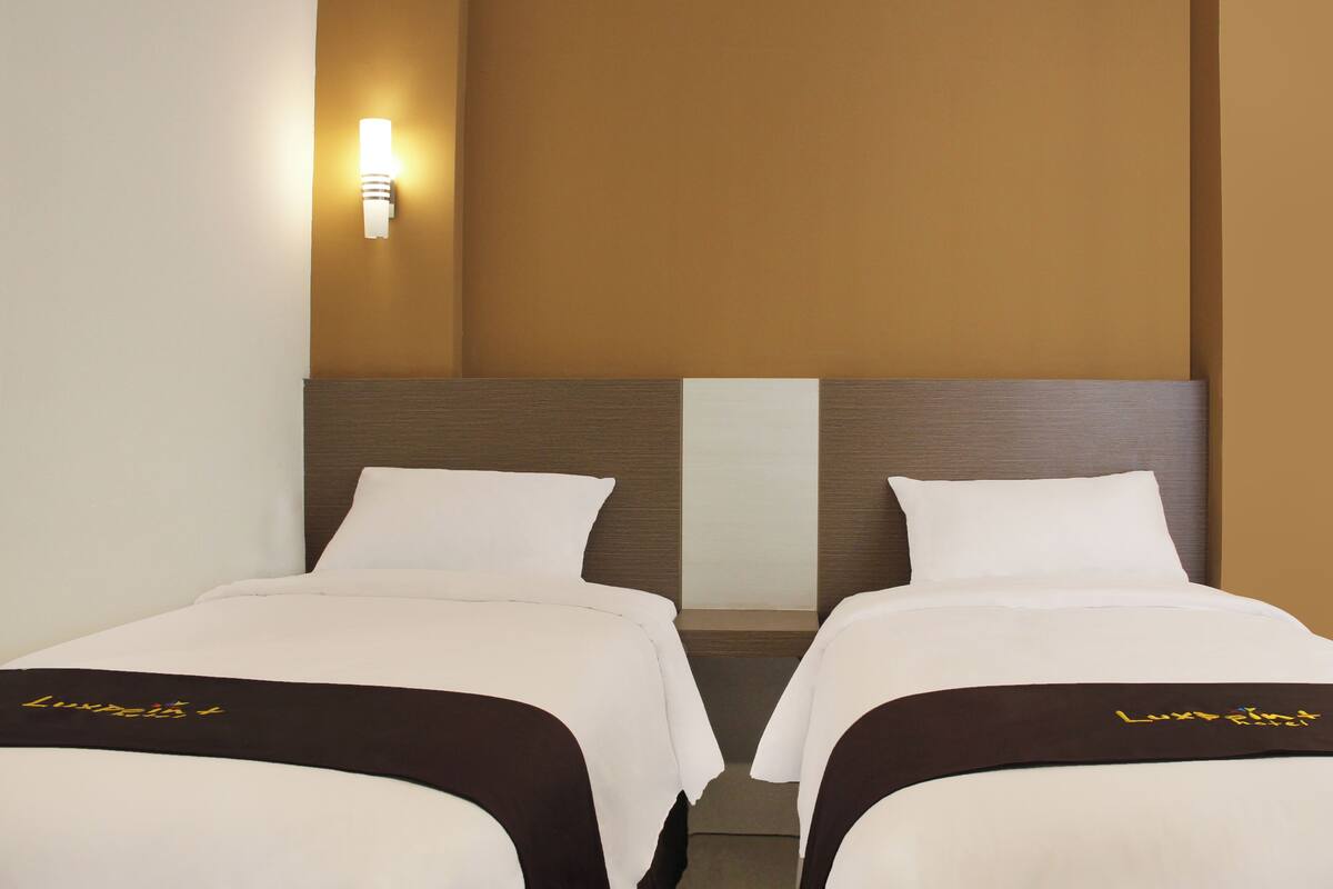 A hotel room with two beds, a nightstand, a wall-mounted light, and a plain wall.