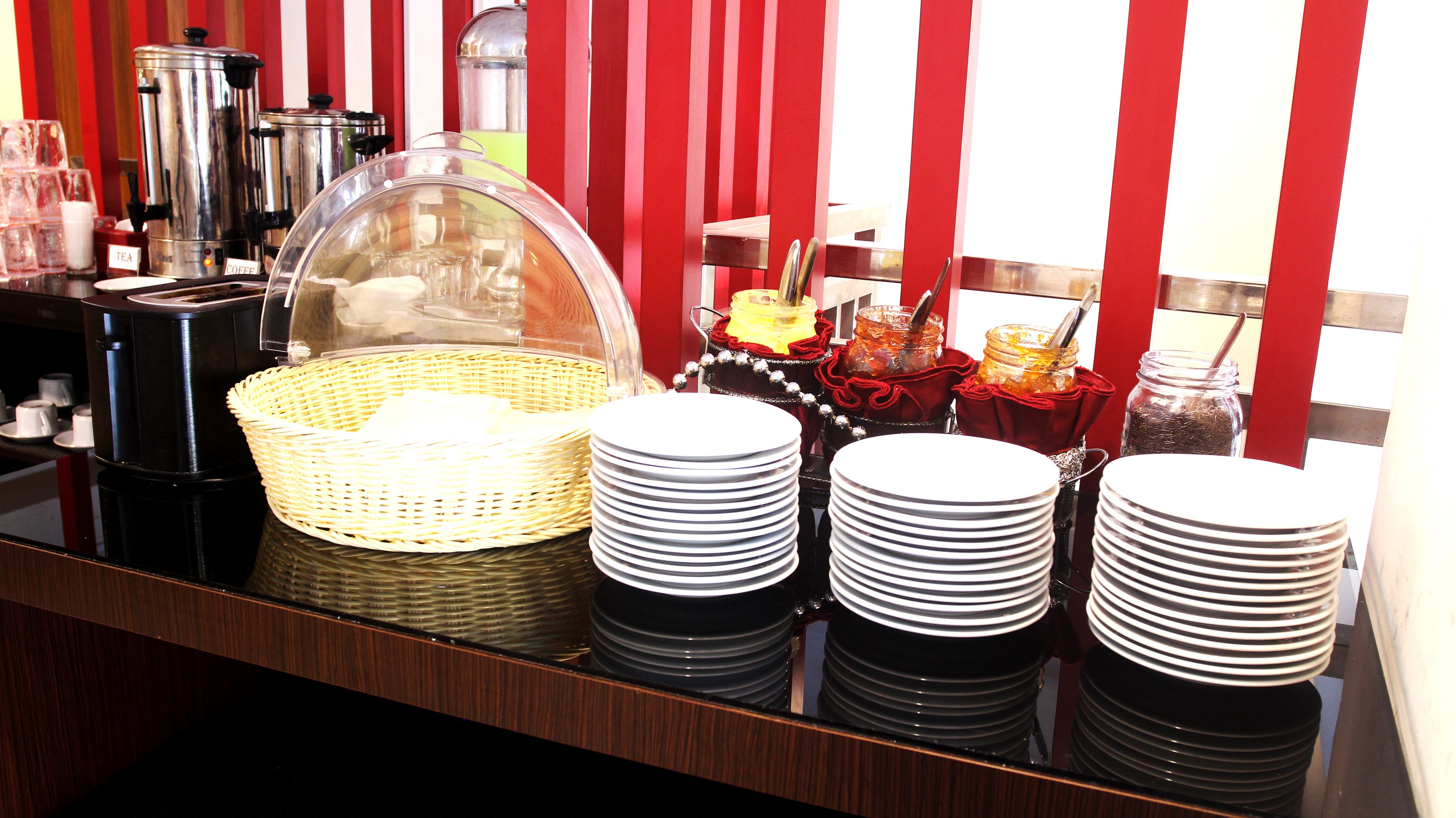 A buffet setup with coffee and tea dispensers, a covered food display, and stacked plates.