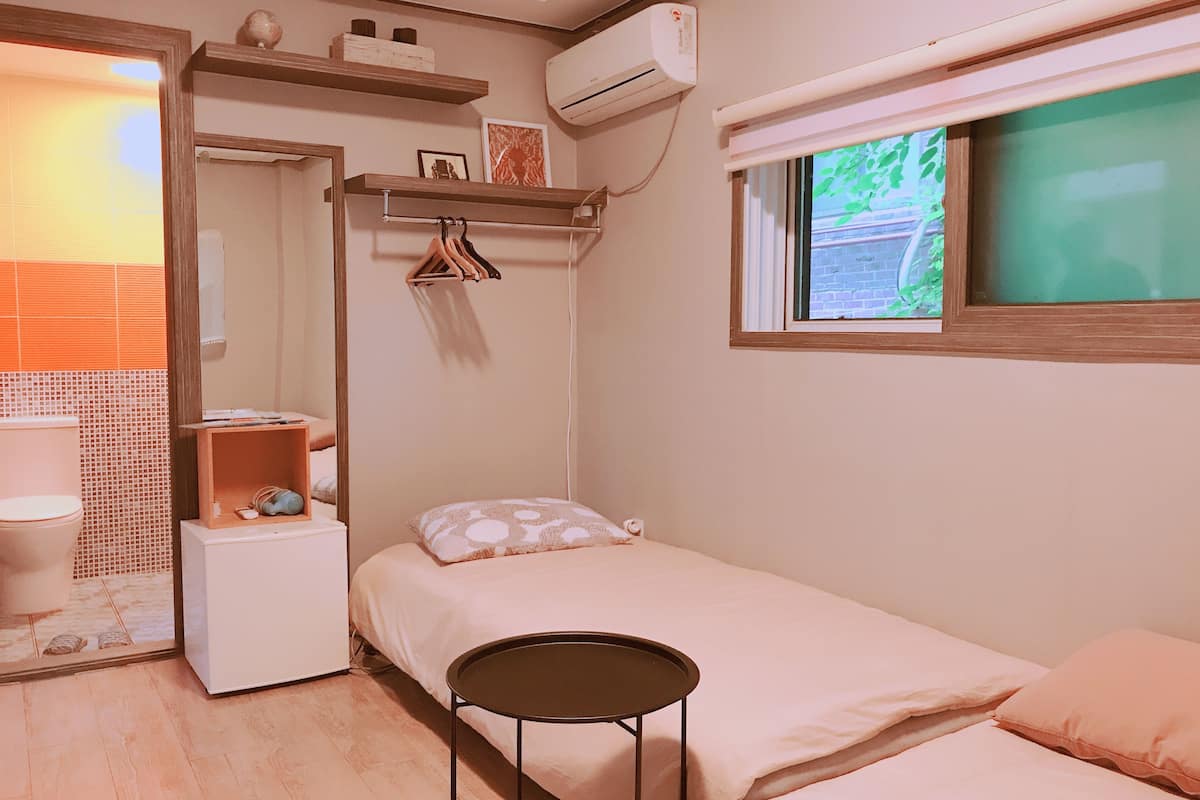 A small, neatly organized bedroom with a single bed, a small table, a wardrobe, and a window with a view of greenery.