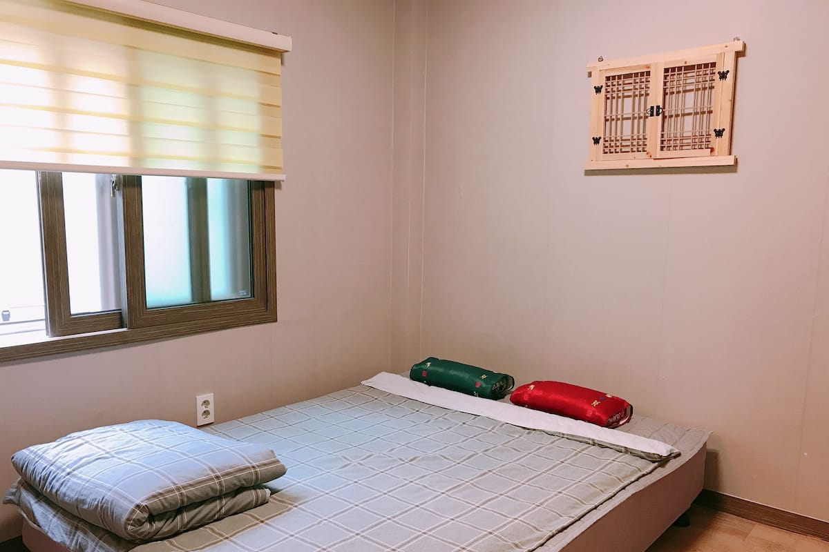 A small, single-bed room with a window, a wall-mounted calendar, and a bed with a checkered bedspread and pillows.