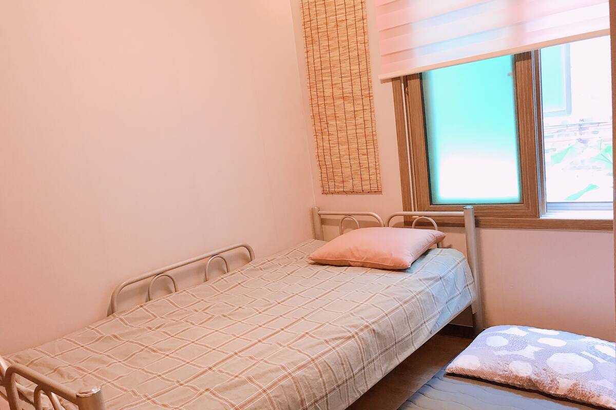 A small, single-bed room with a metal frame, a window with blinds, and a wall-mounted woven curtain.