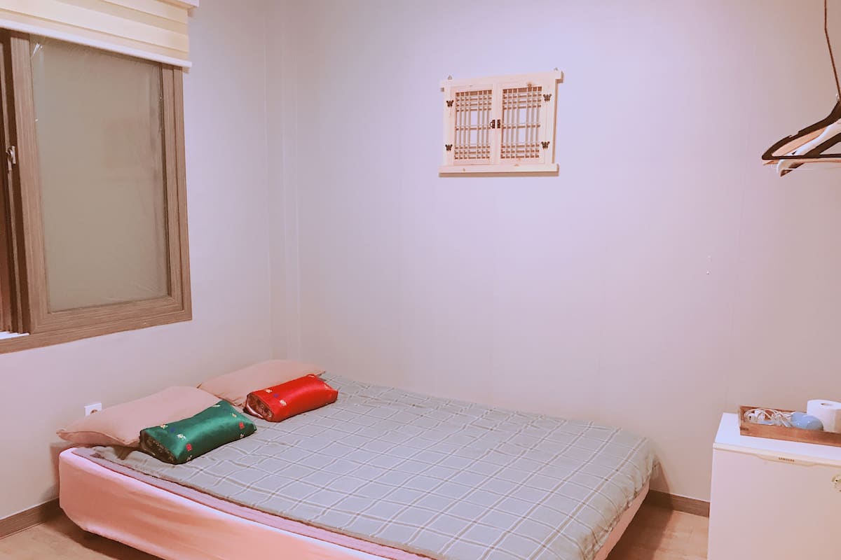 A small bedroom with a bed, a window, a wall-mounted heater, and a small white dresser.