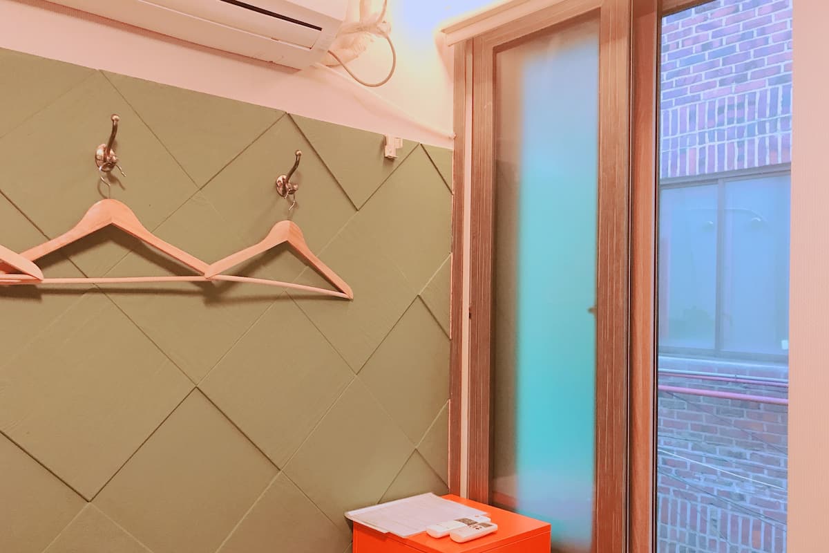 A room with a green wall, wooden door, and a window with a view of a brick building. There are two wooden hangers on the wall and a small orange table with a white object on it.