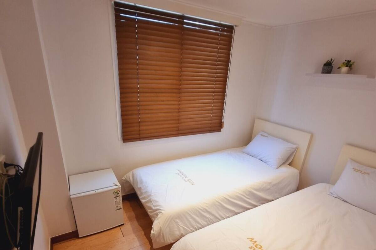 Una habitación pequeña y limpia con dos camas, una ventana con persianas, una mesita de noche y una lámpara en el techo.