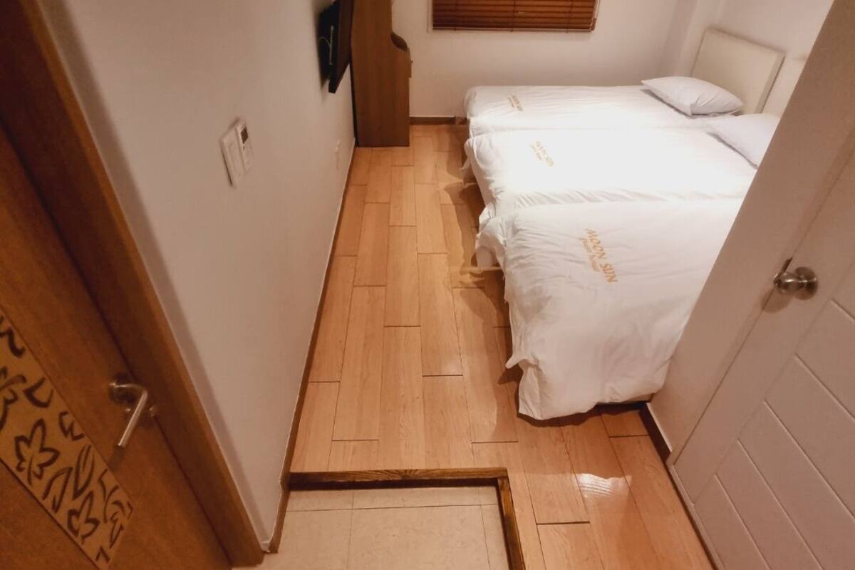 Un pasillo estrecho que lleva a una habitación con dos camas, suelo de madera y una ventana con persianas.