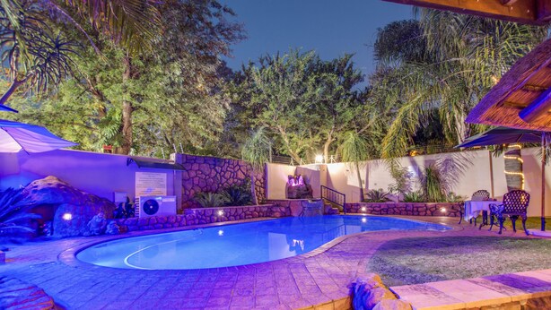 A swimming pool with a stone border, surrounded by tropical plants and trees, and a stone wall with a sign. - Kassaboera Lodge (Hartbeespoort)