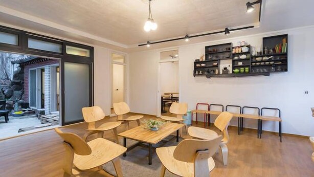 A modern interior with a round wooden table surrounded by chairs, a bookshelf, and a view through a glass door. - 9 Road - Hostel (Seoul)