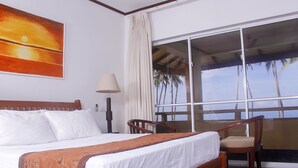 Une chambre d’hôtel avec un lit, une chaise, une lampe et une grande fenêtre donnant sur des palmiers et l’océan.