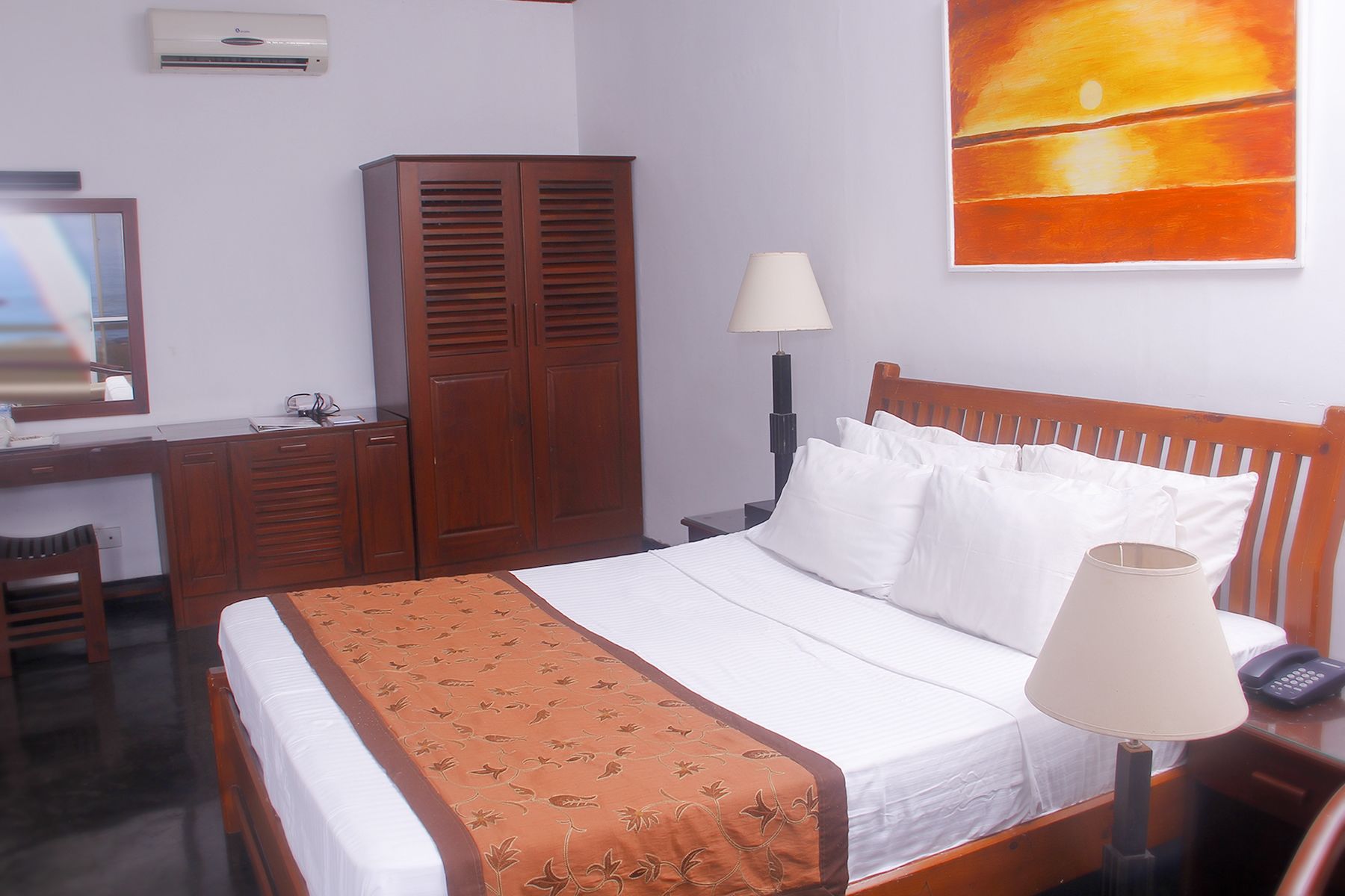 Quarto de hotel com cama, escrivaninha, guarda-roupa, abajur e um quadro na parede.