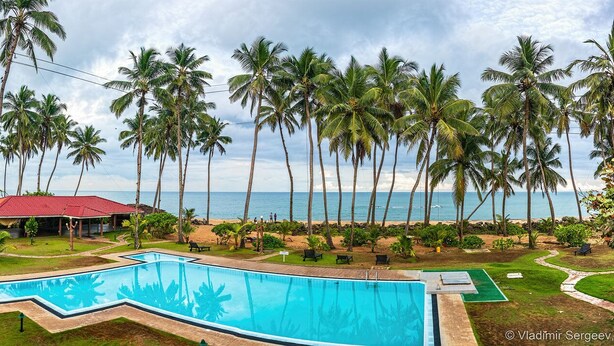 Un espace piscine avec un bâtiment à toit rouge, des palmiers et une vue sur la plage et l’océan.