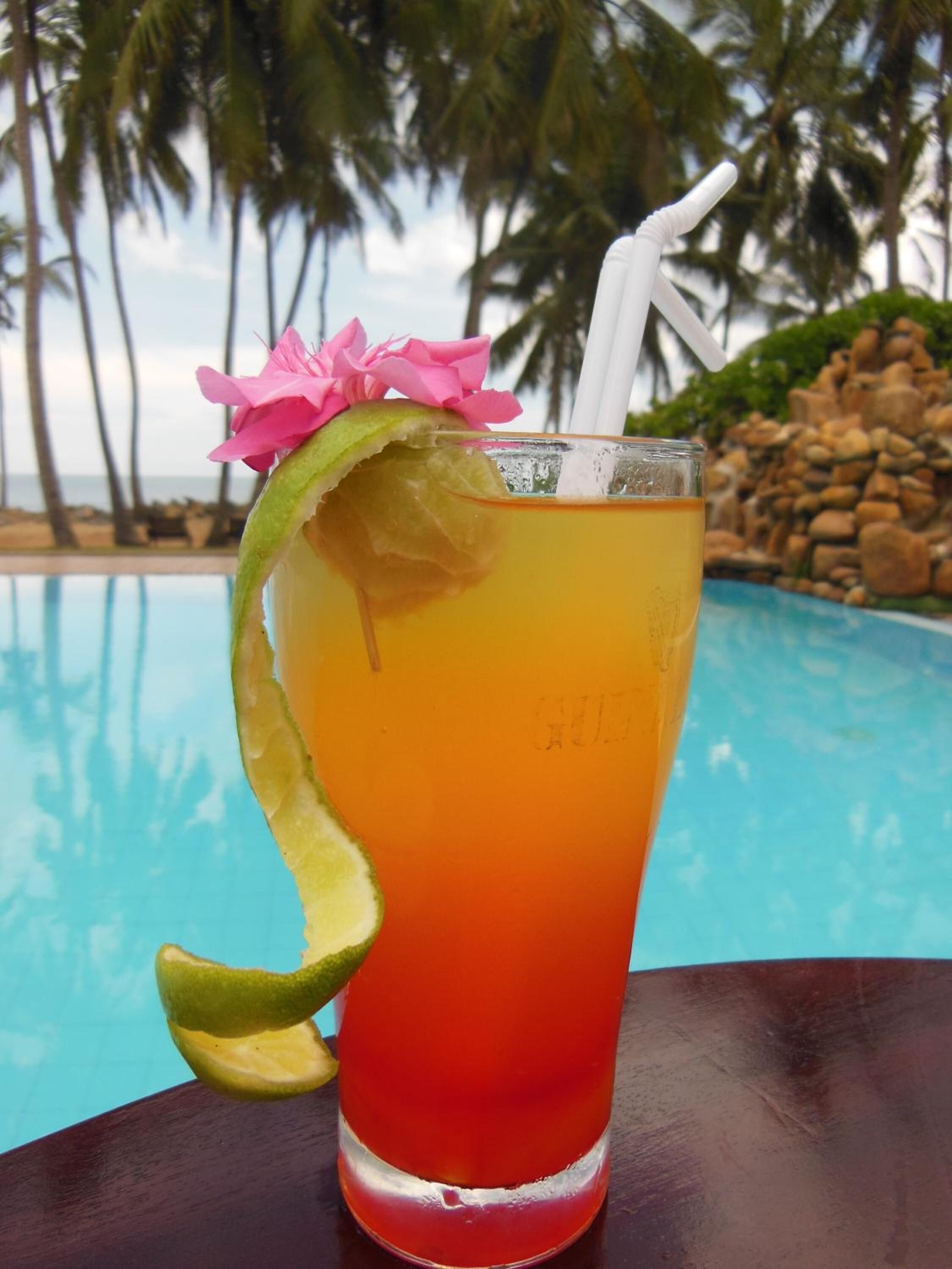 A tropical drink with a lime wedge and a pink flower garnish, placed on a table by a pool with palm trees and a rocky shoreline in the background.