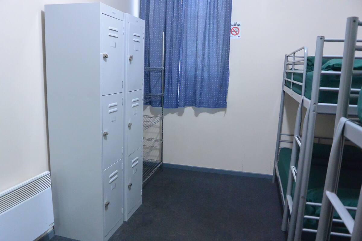 Une petite pièce avec un lit superposé, une armoire en métal et une fenêtre avec des rideaux.