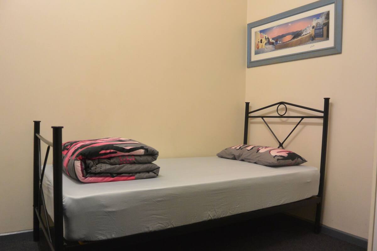 Un lit simple avec un cadre en métal, un matelas gris et une couverture rose et grise.