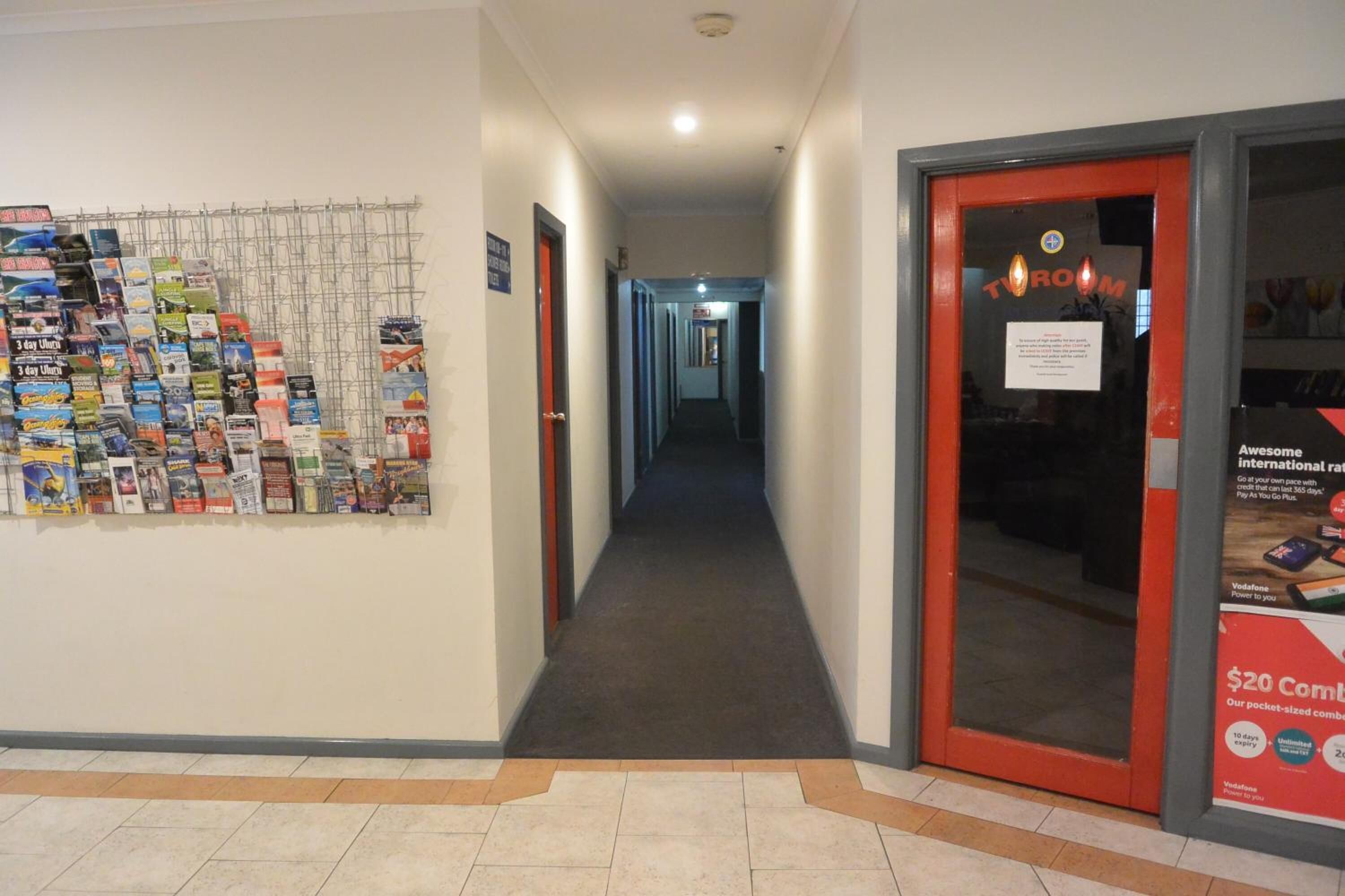 A hallway with a display of travel brochures on the left, a red door with a sign on the right, and a tiled floor.
