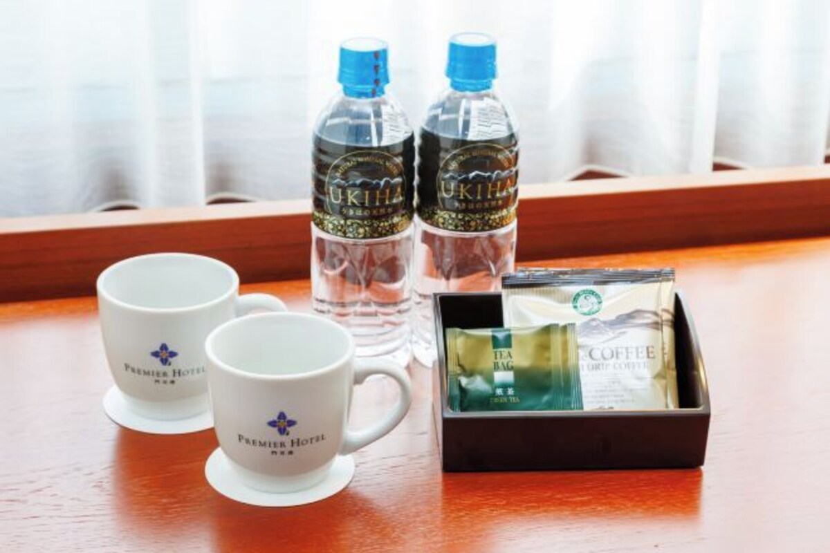Dos tazas blancas con el logotipo de "Premier Hotel", dos botellas de agua y una caja con té y café.