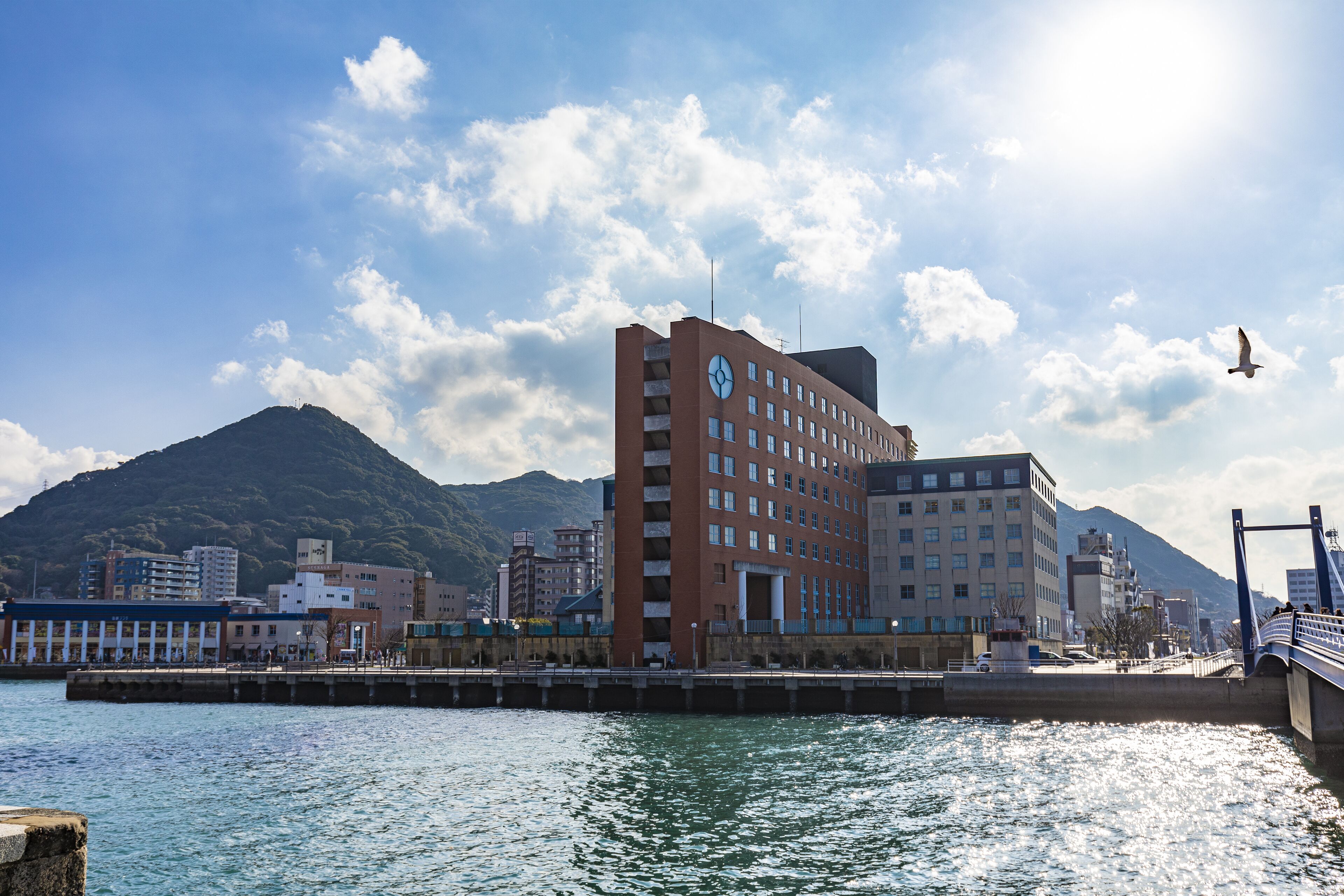 A waterfront hotel with a clock tower, a bridge, and mountains in the background.