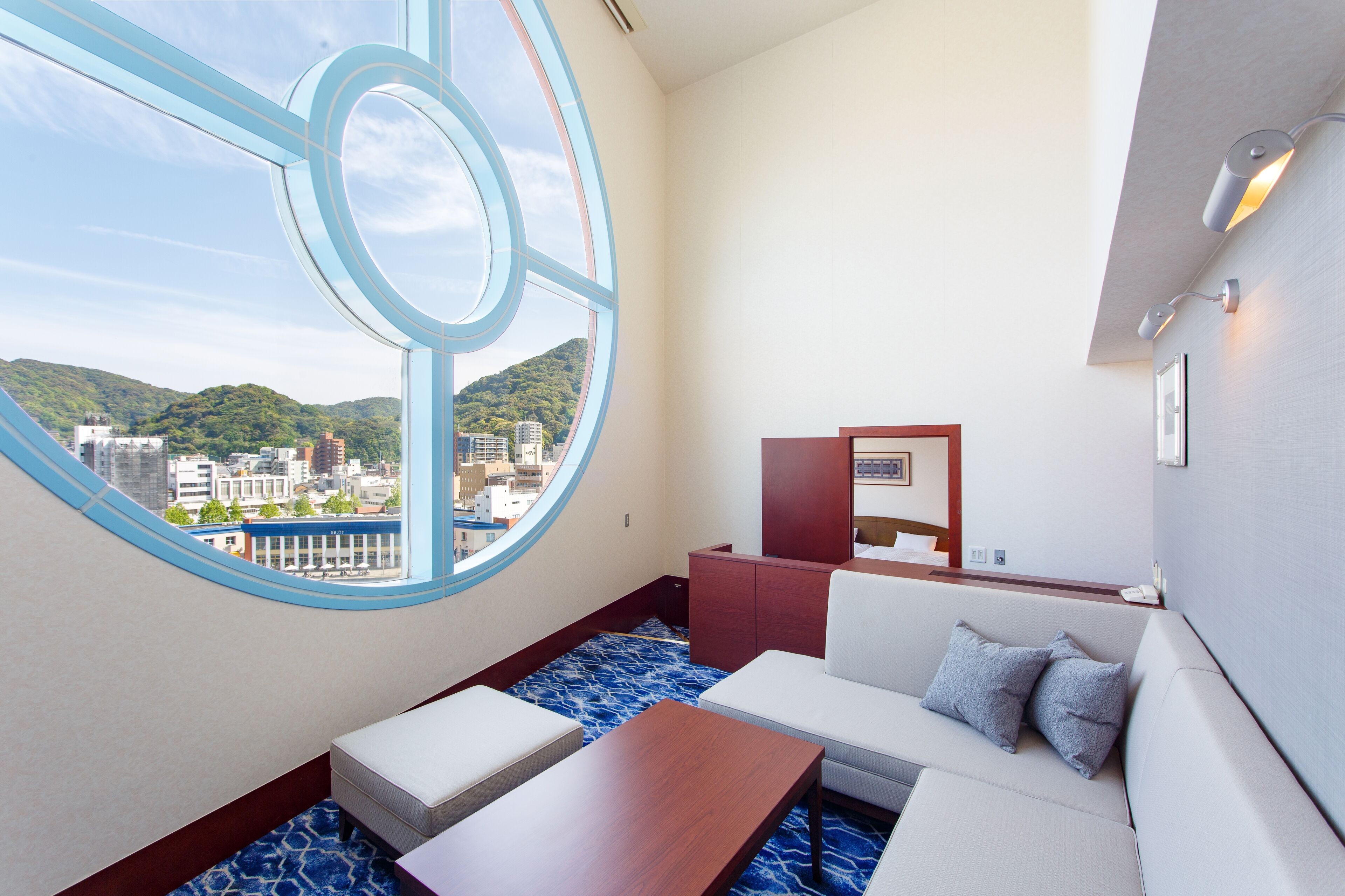 A modern hotel room with a large window offering a city view, a sofa, a small table, and a cabinet.