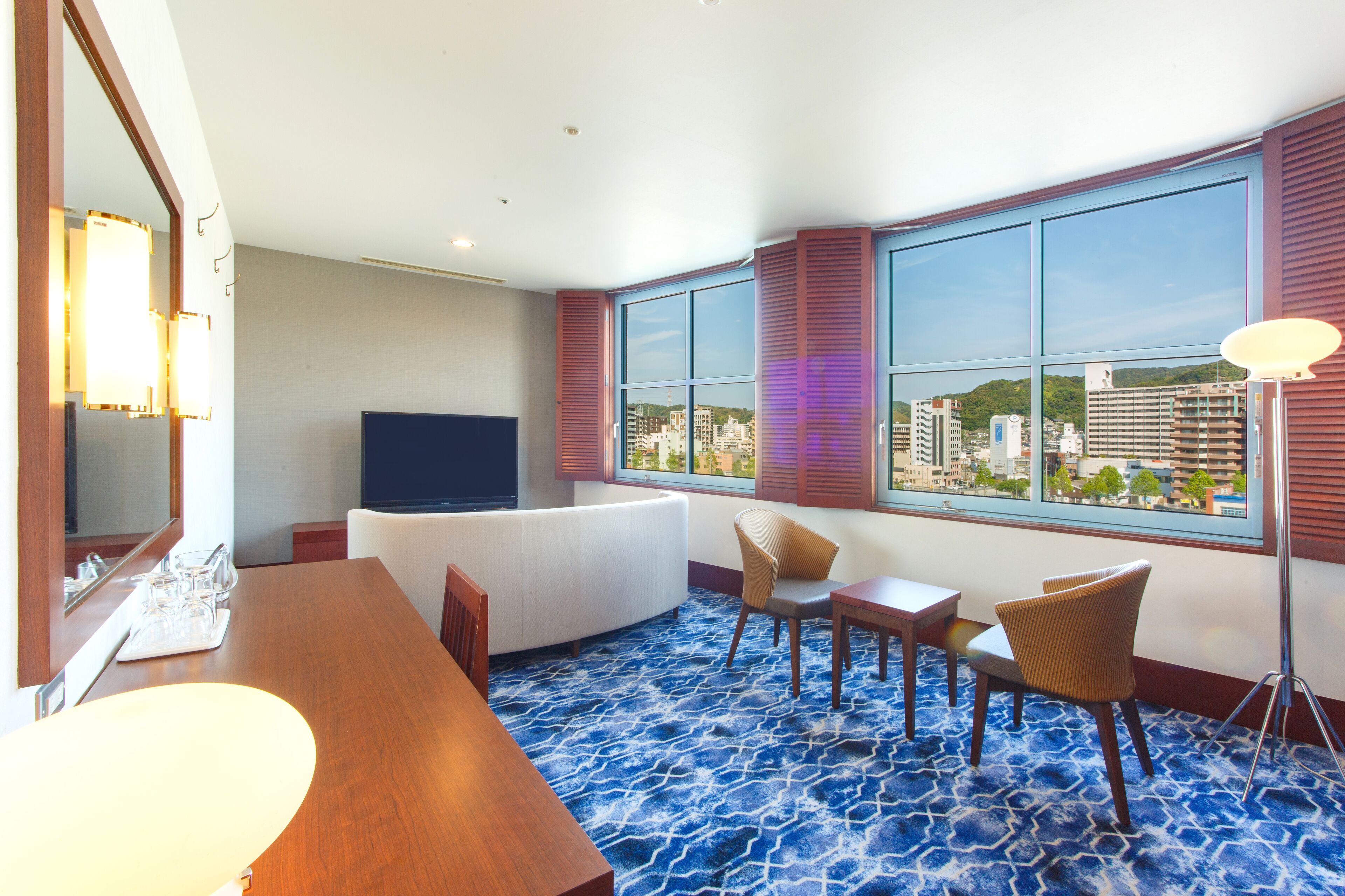 A hotel room with a large window, a flat-screen TV, a dining table, and a seating area with chairs.