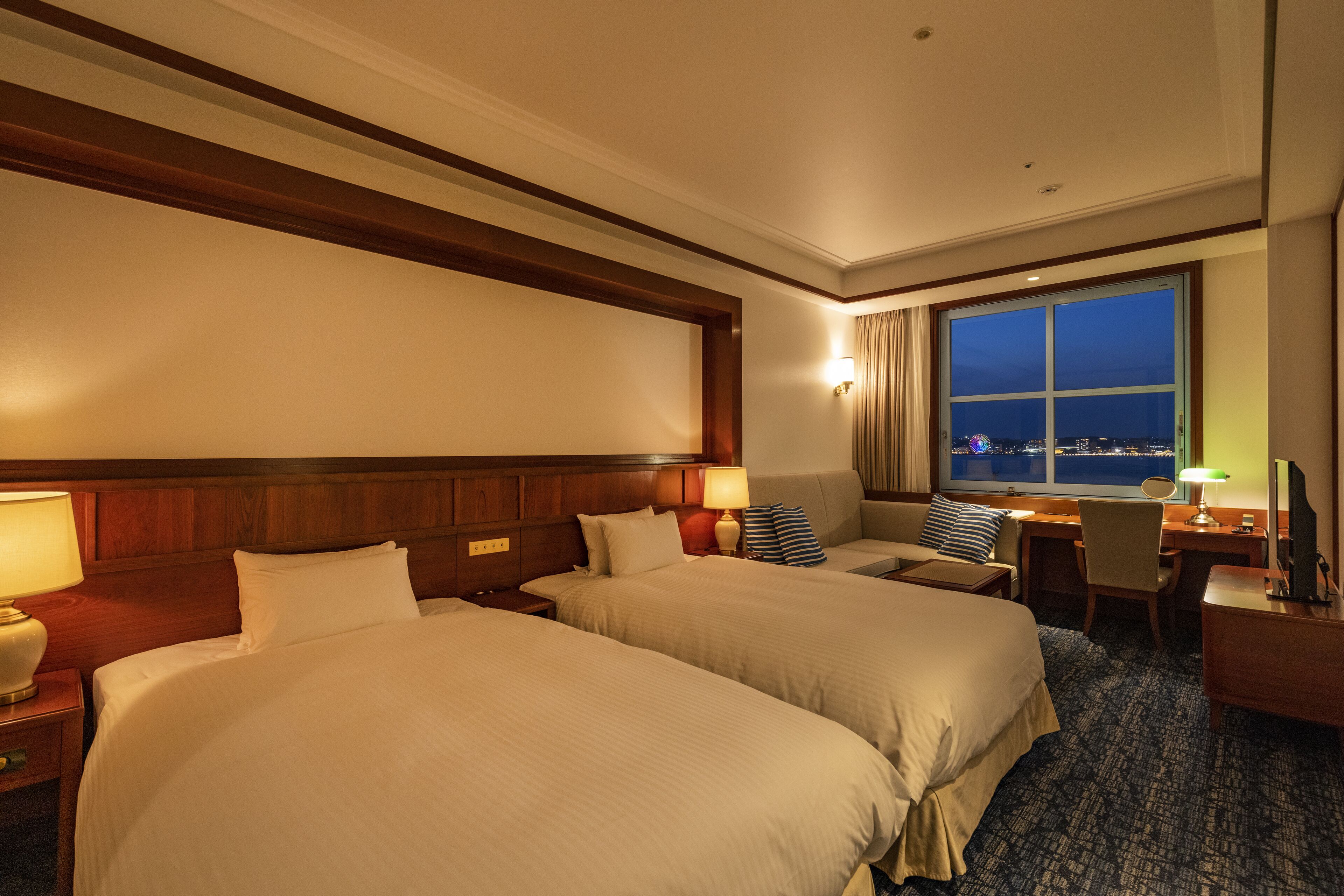 A hotel room with two beds, a desk, a chair, a window with a city view, and a nightstand with a lamp.