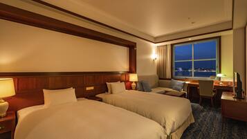 A hotel room with two beds, a desk, a chair, a window with a city view, and a nightstand with a lamp.