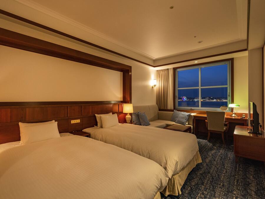 A hotel room with two beds, a desk, a chair, a window with a city view, and a nightstand with a lamp.