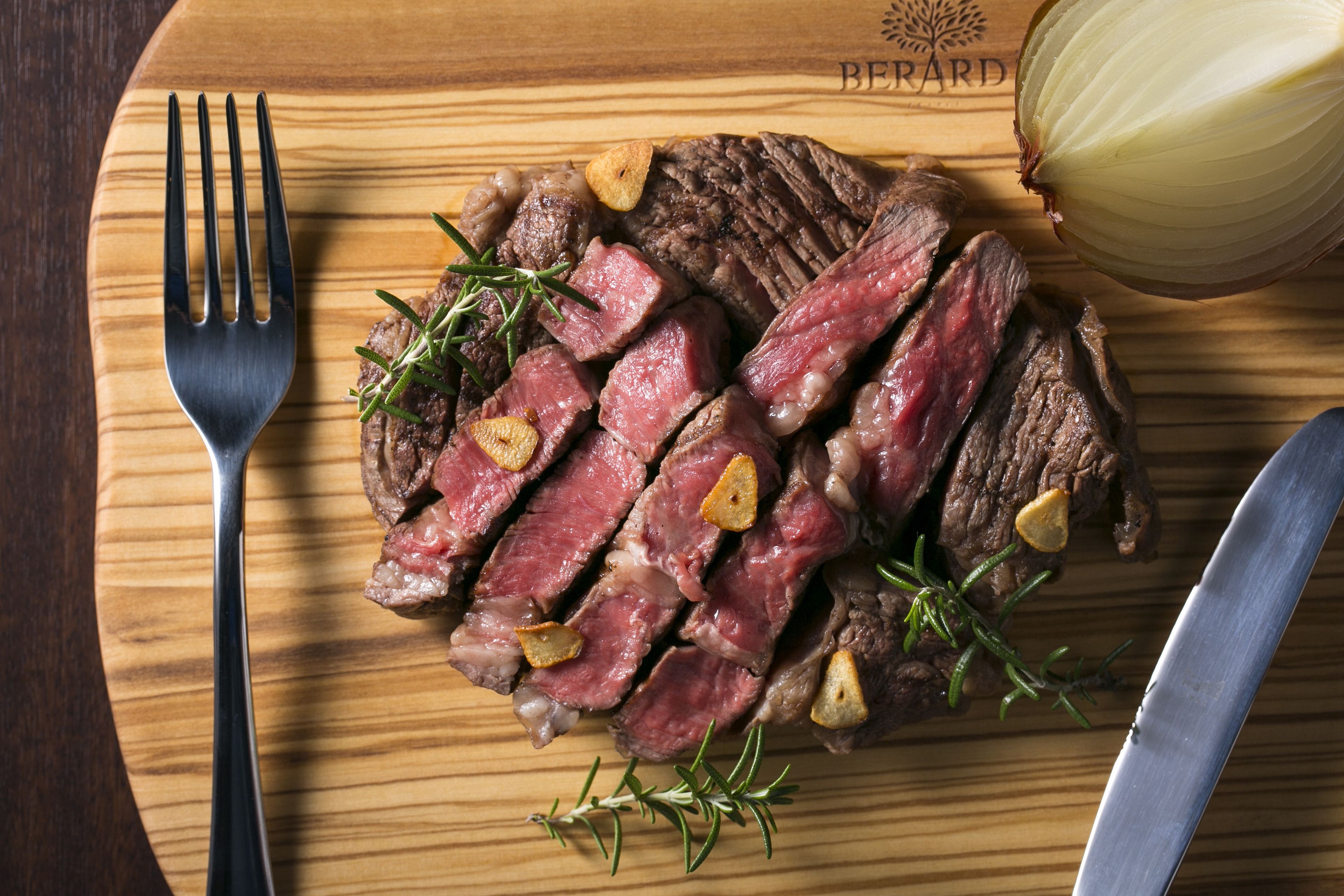 A wooden cutting board with sliced steak, rosemary, and garlic cloves.