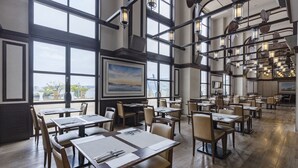 A spacious restaurant with high ceilings, large windows, and a view of a bridge. Tables are neatly arranged with menus and cutlery.