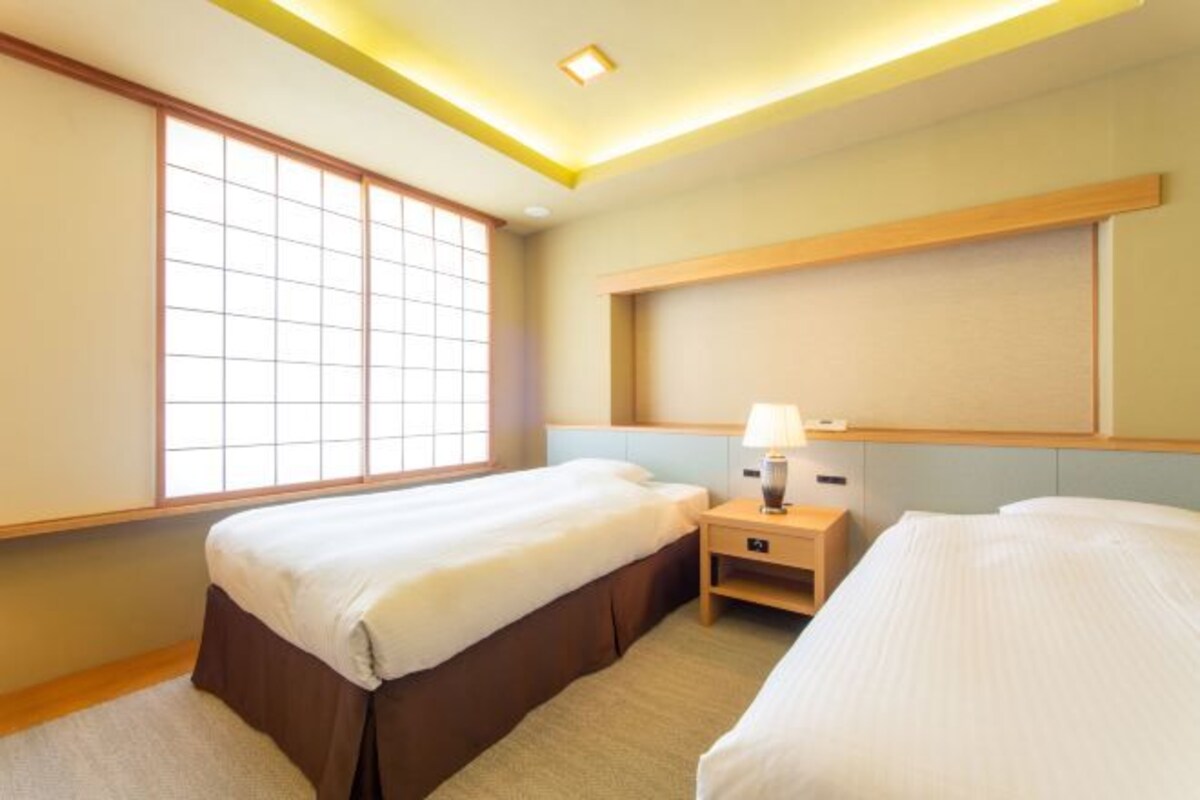 Habitación de hotel con dos camas, una ventana, una mesita de noche con lámpara e iluminación en el techo.