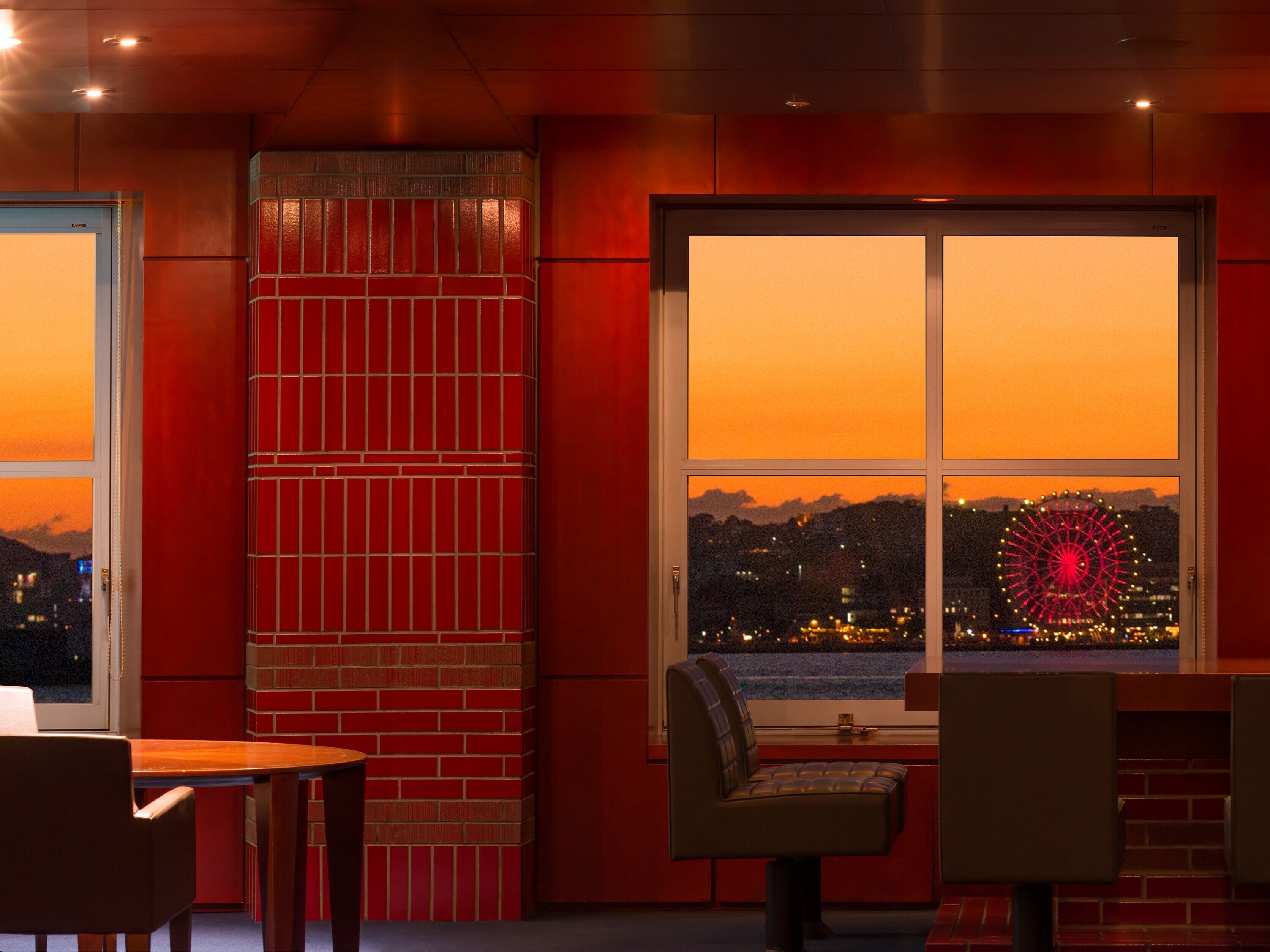 A room with a brick wall, a large window with a view of a cityscape and a Ferris wheel, and a seating area with a table and chairs.