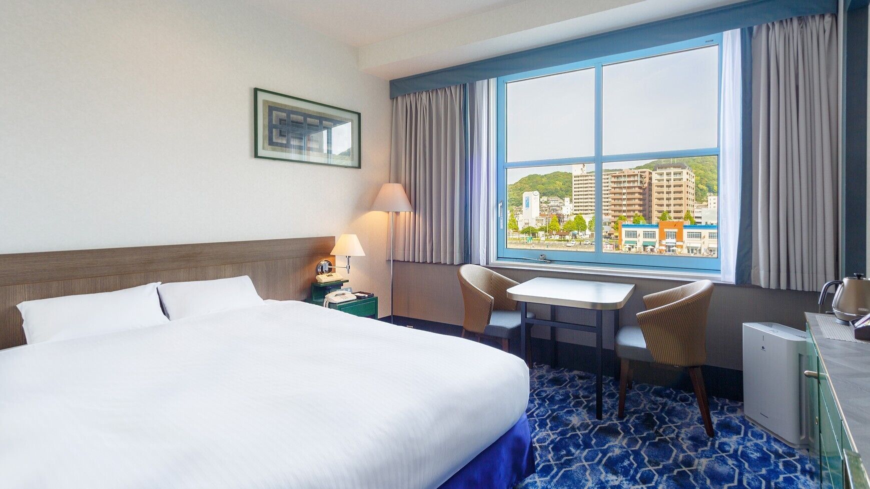 Quarto de hotel com uma cama grande, uma escrivaninha, duas cadeiras, uma janela com vista para a cidade e uma pequena mesa com uma chaleira.