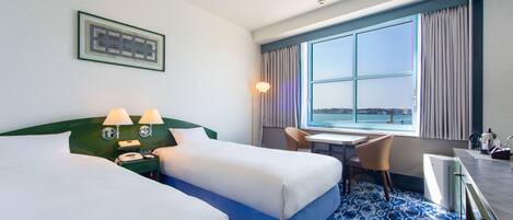 A hotel room with two beds, a desk, a chair, a view of the sea, and a window with curtains.