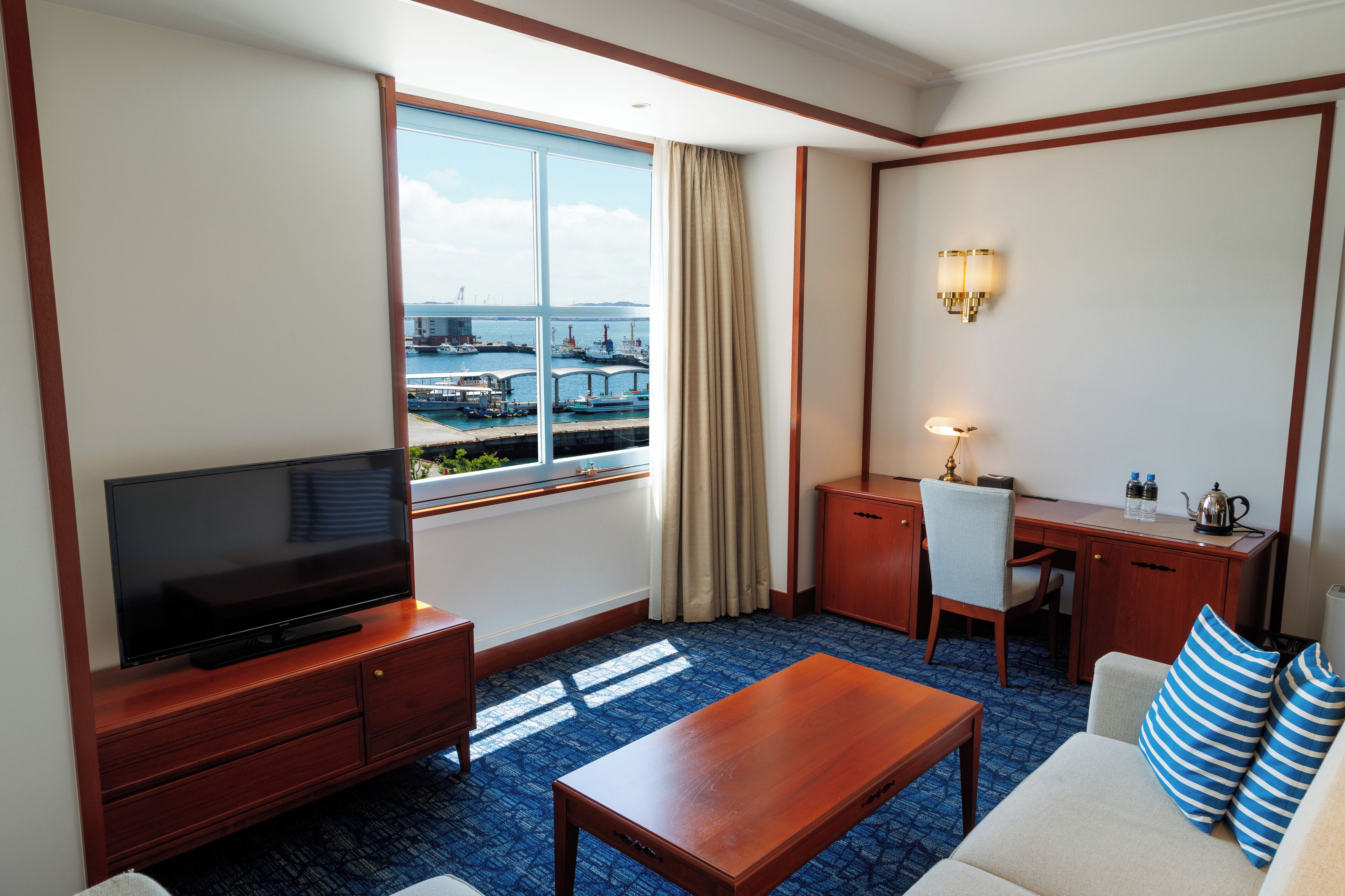 A hotel room with a television, a desk, a sofa, and a view of a marina.
