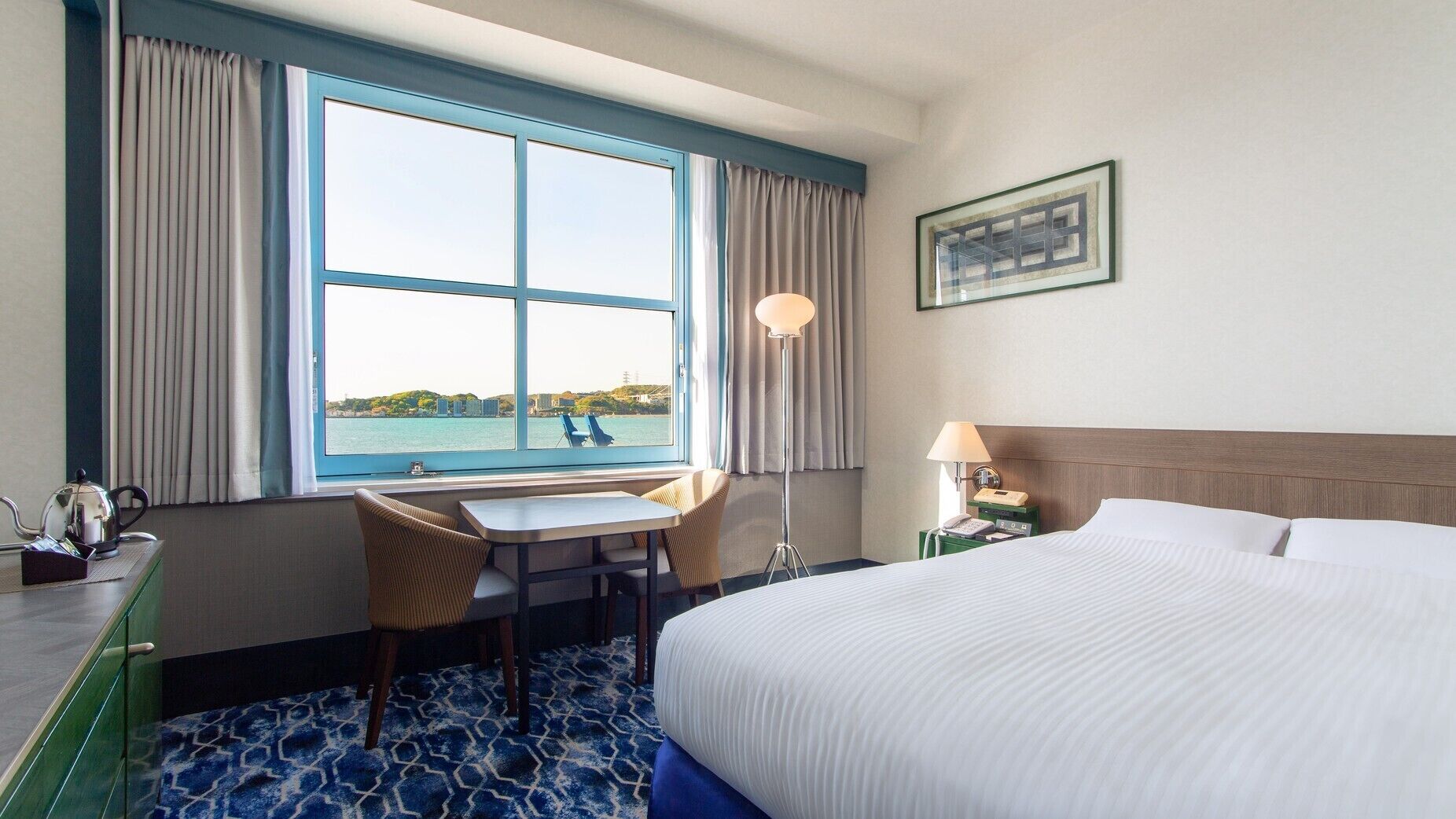 Quarto de hotel com cama, escrivaninha com cadeiras, luminária, mesa de cabeceira e vista para um corpo d'água.