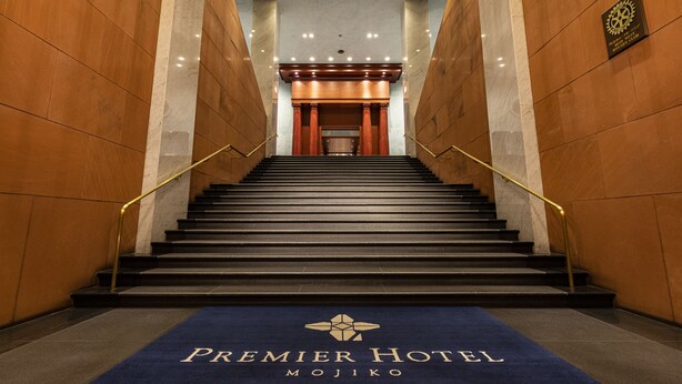 A hotel lobby with a marble floor, a blue carpet with "Premier Hotel Mojiko" text, and a staircase with a handrail.