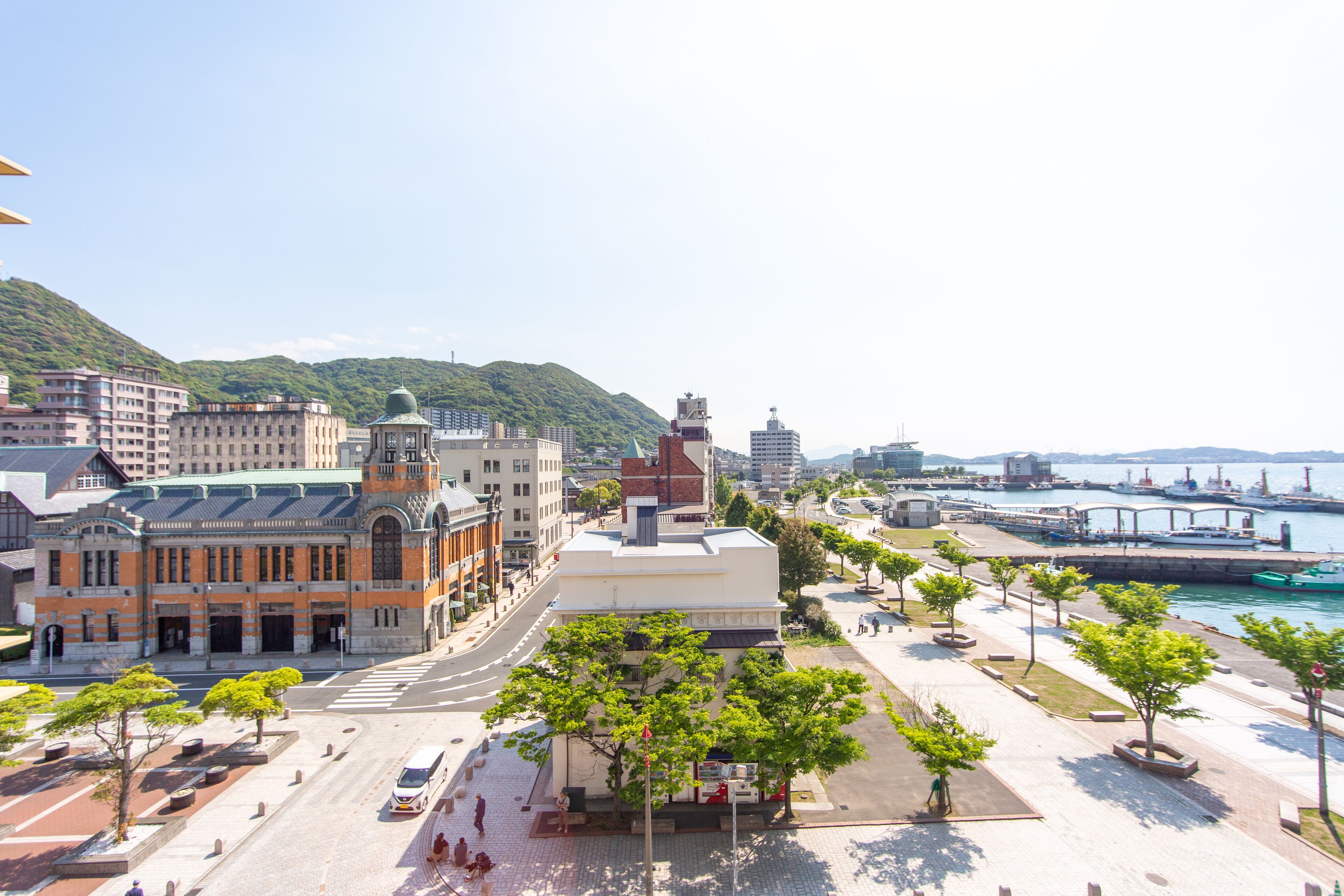 A cityscape with a prominent building featuring a green roof and a dome, a waterfront with a bridge, and a pedestrian area with trees and benches.
