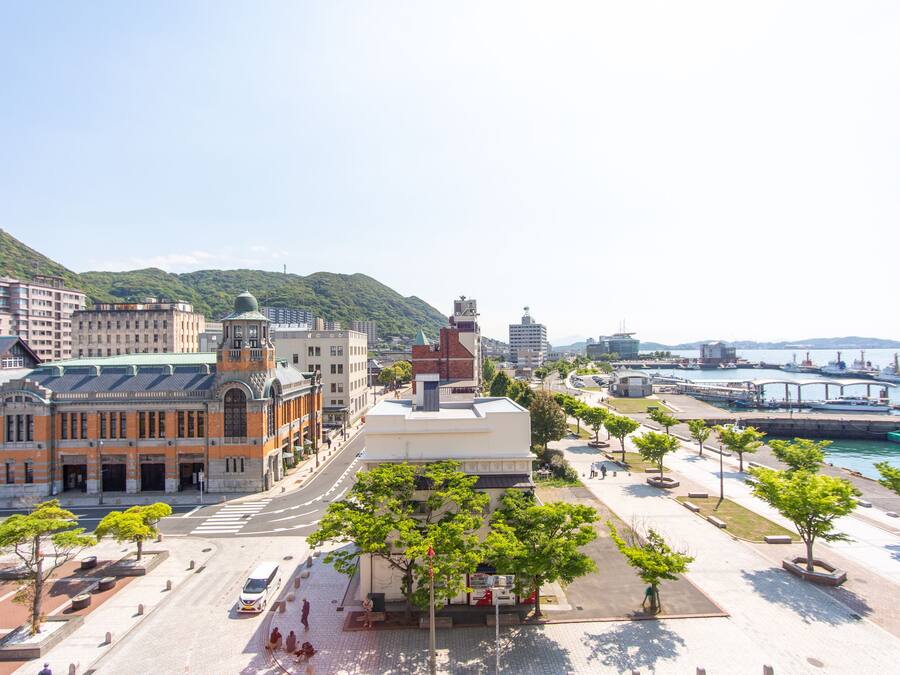 A cityscape with a prominent building featuring a green roof and a dome, a waterfront with a bridge, and a pedestrian area with trees and benches.